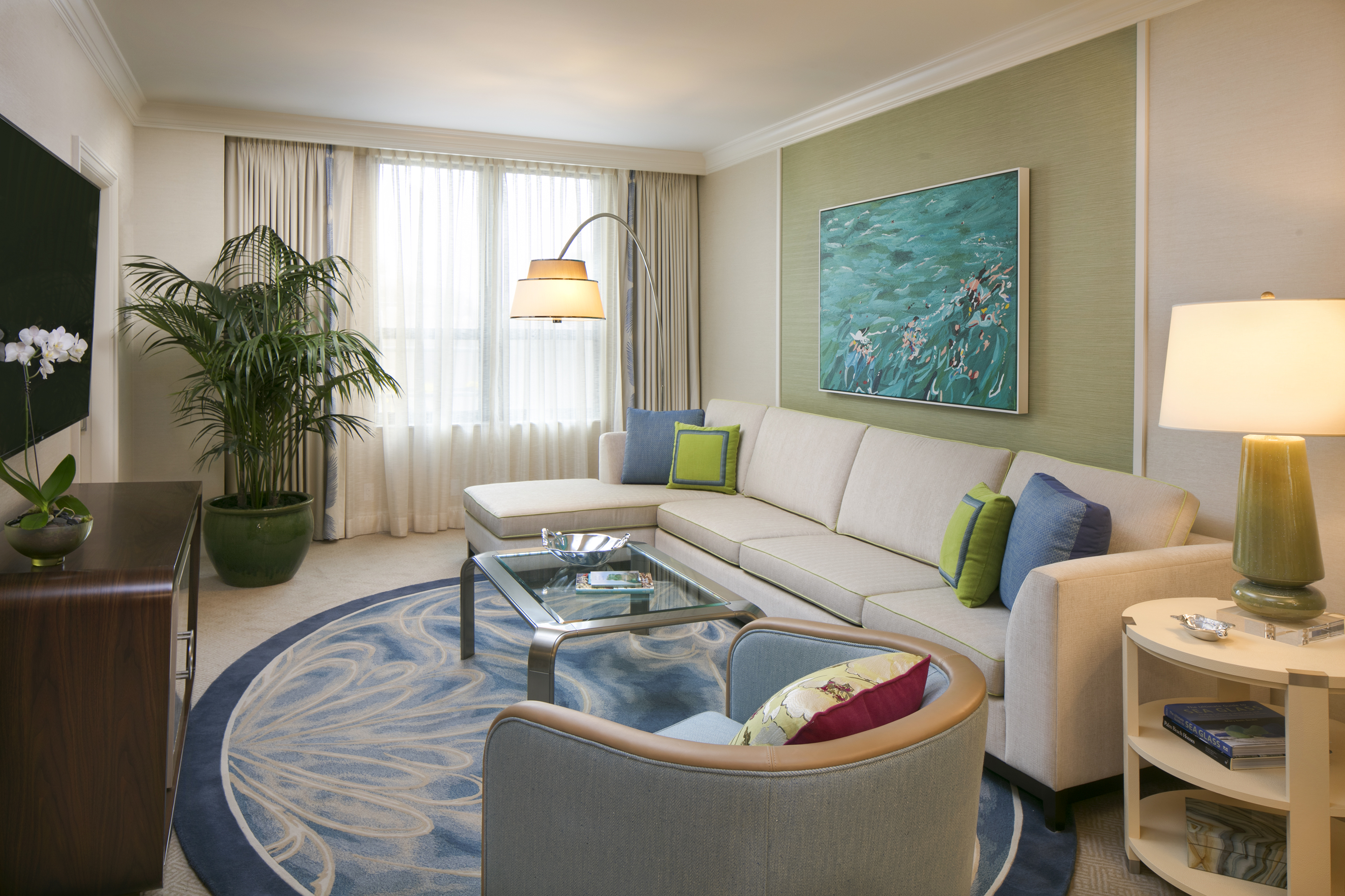 Modern living room with beige sectional sofa, blue patterned rug, ocean artwork, and green accents.