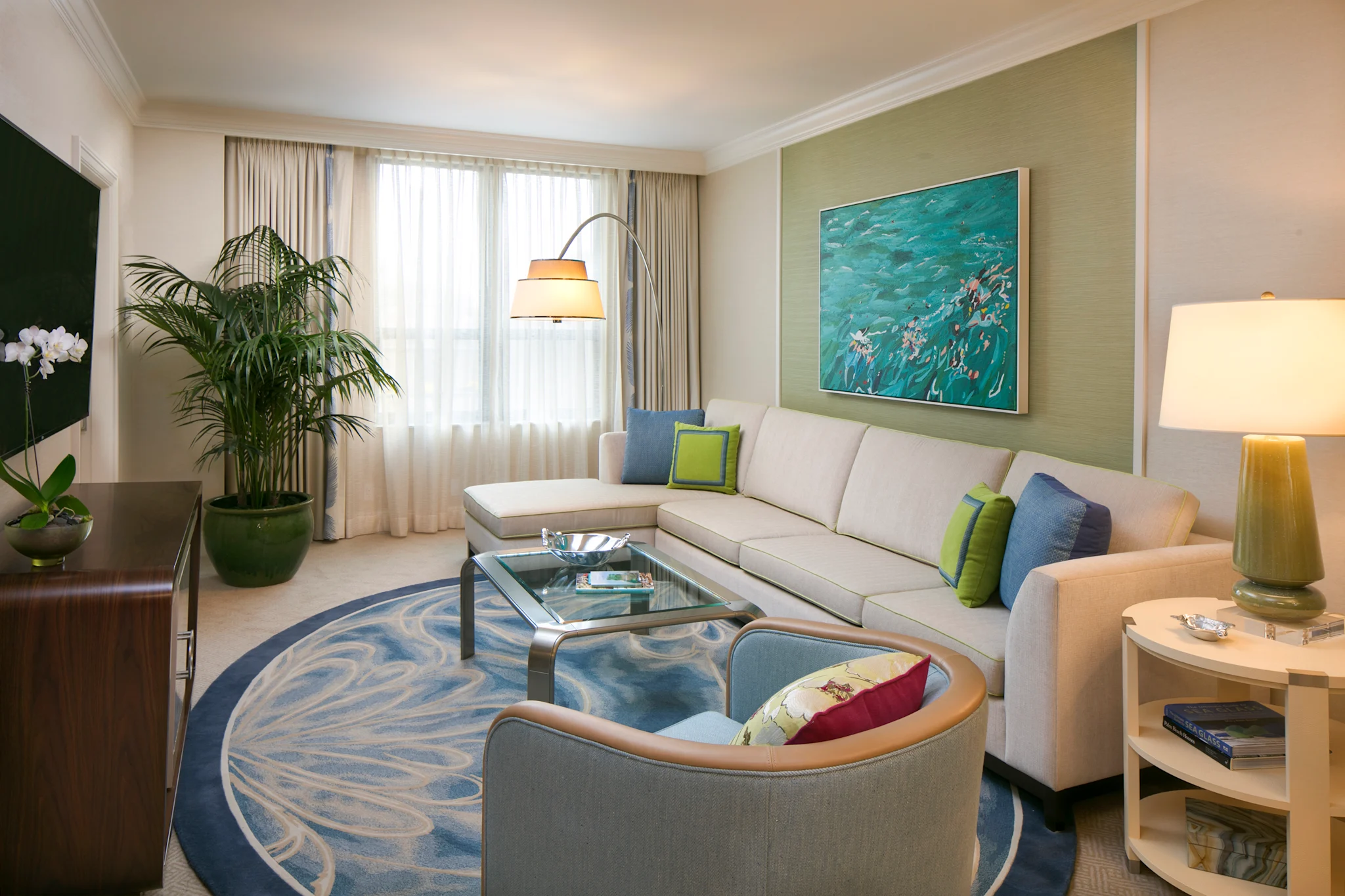 Modern living room with beige sectional sofa, blue patterned rug, ocean artwork, and green accents.
