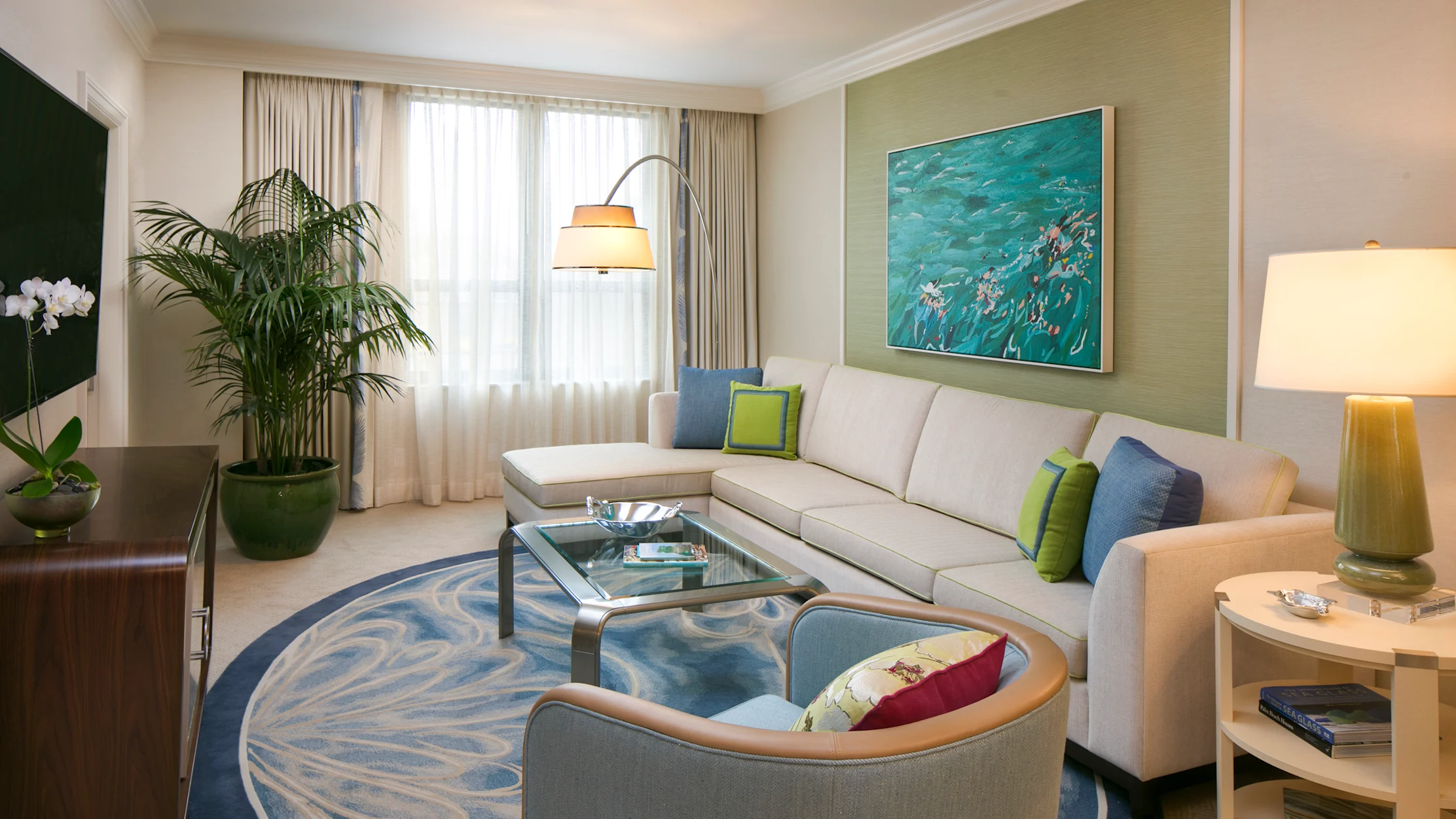 Modern living room with beige sectional sofa, blue patterned rug, ocean artwork, and green accents.