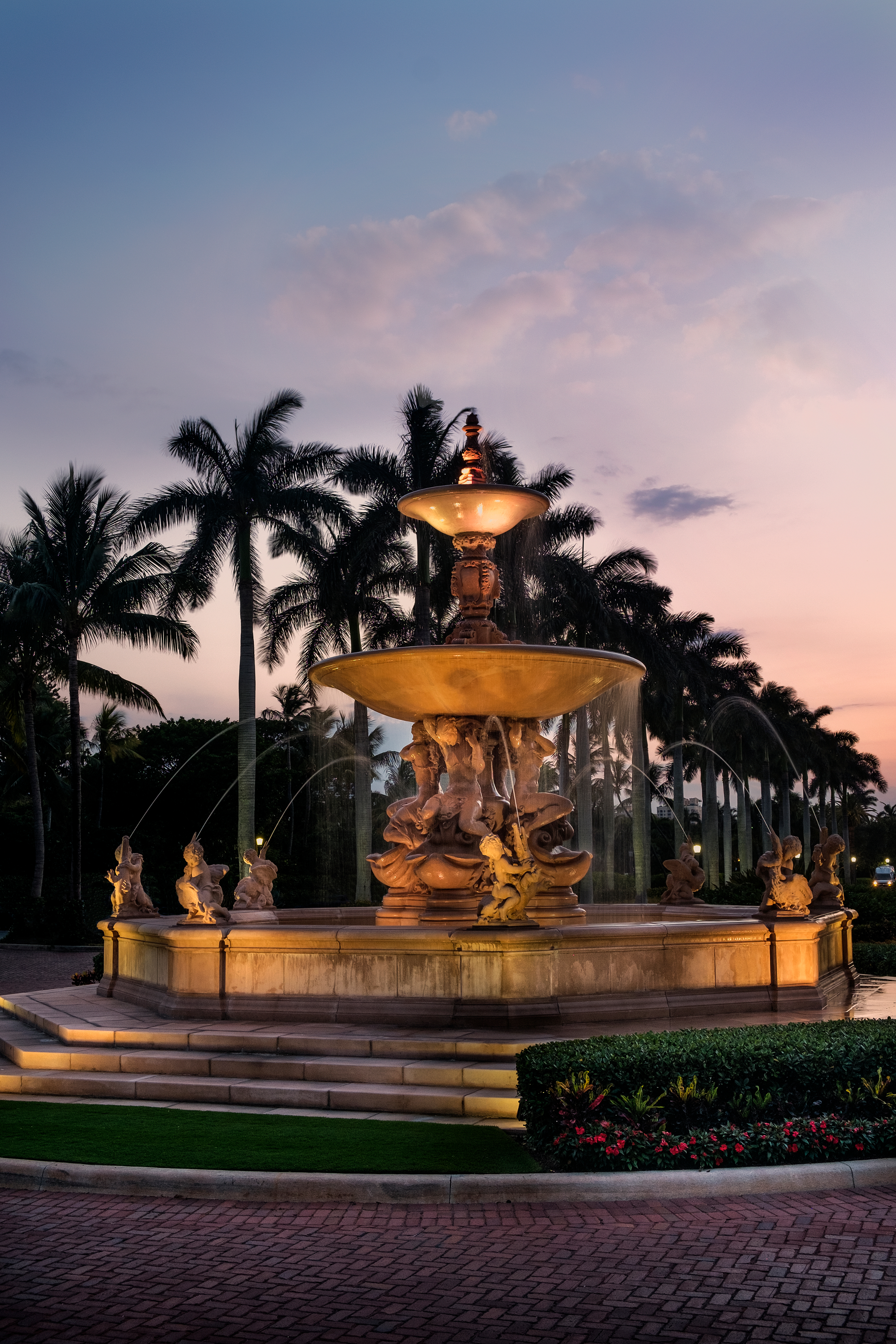 Illuminated ornate fountain with tiered basins and statues at dusk, palm trees silhouetted behind.
