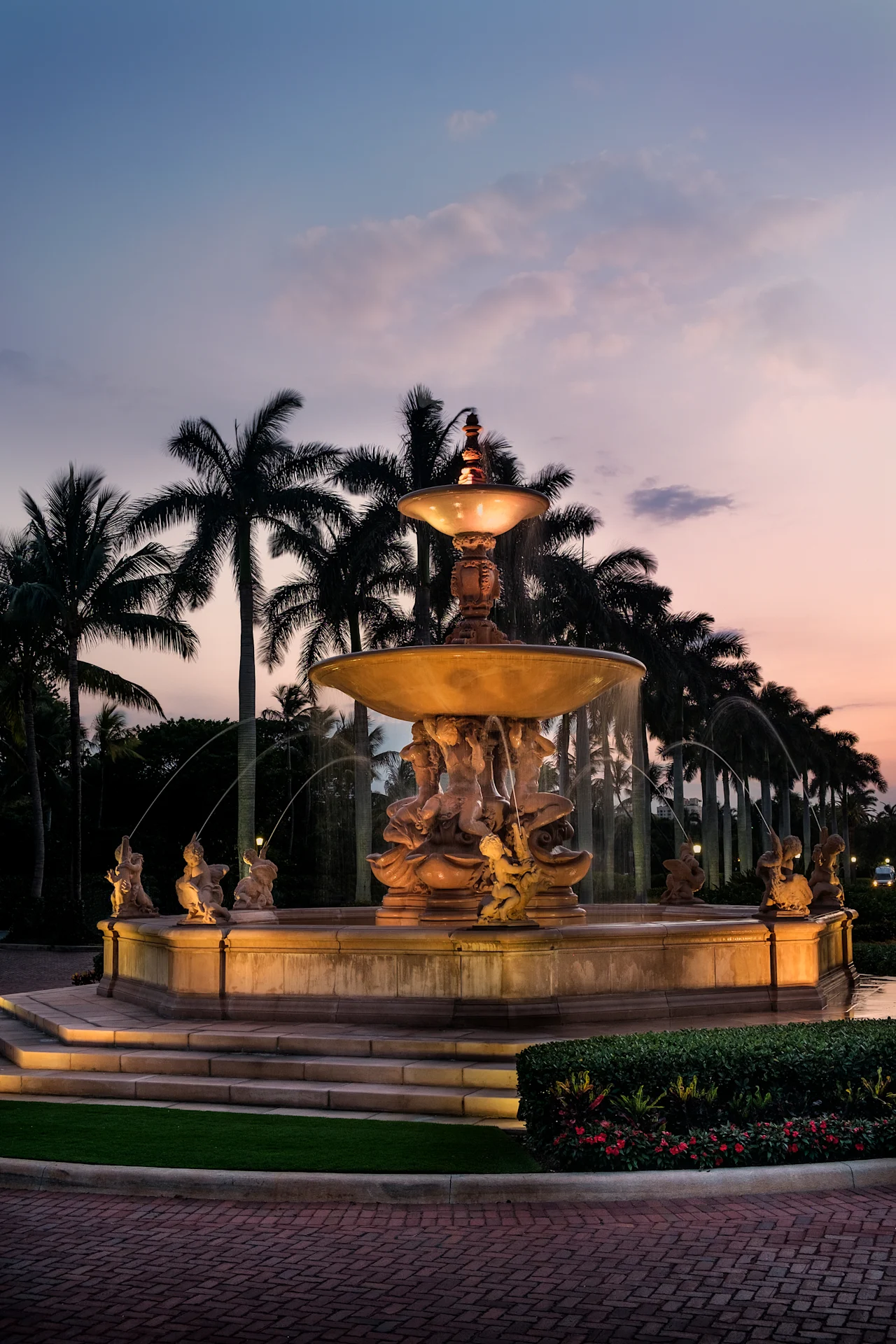 Illuminated ornate fountain with tiered basins and statues at dusk, palm trees silhouetted behind.