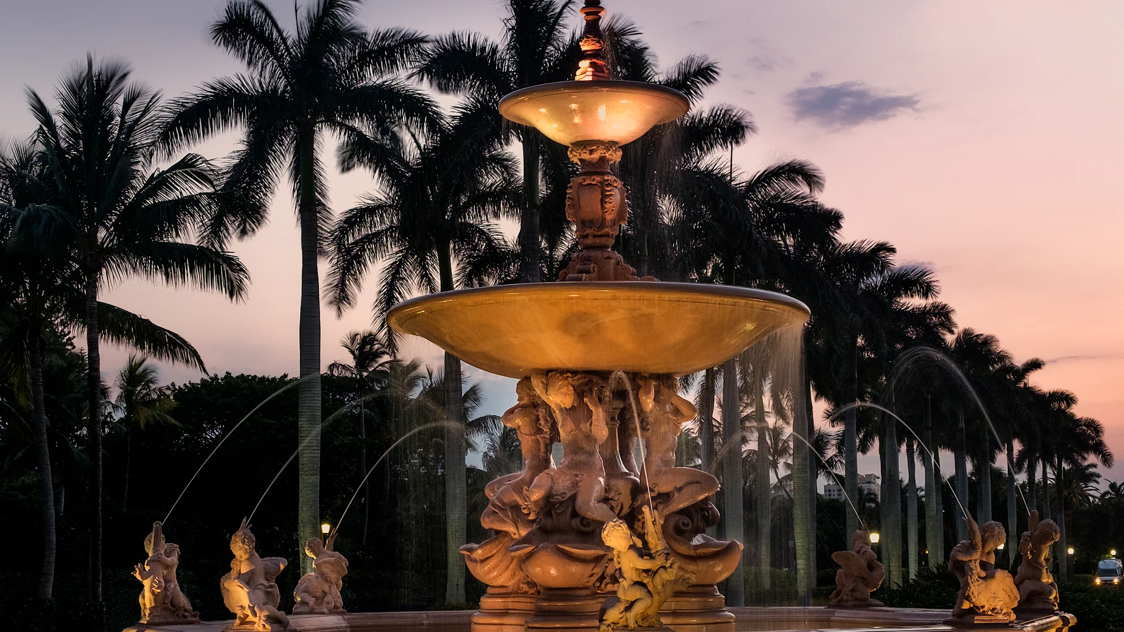 Illuminated ornate fountain with tiered basins and statues at dusk, palm trees silhouetted behind.