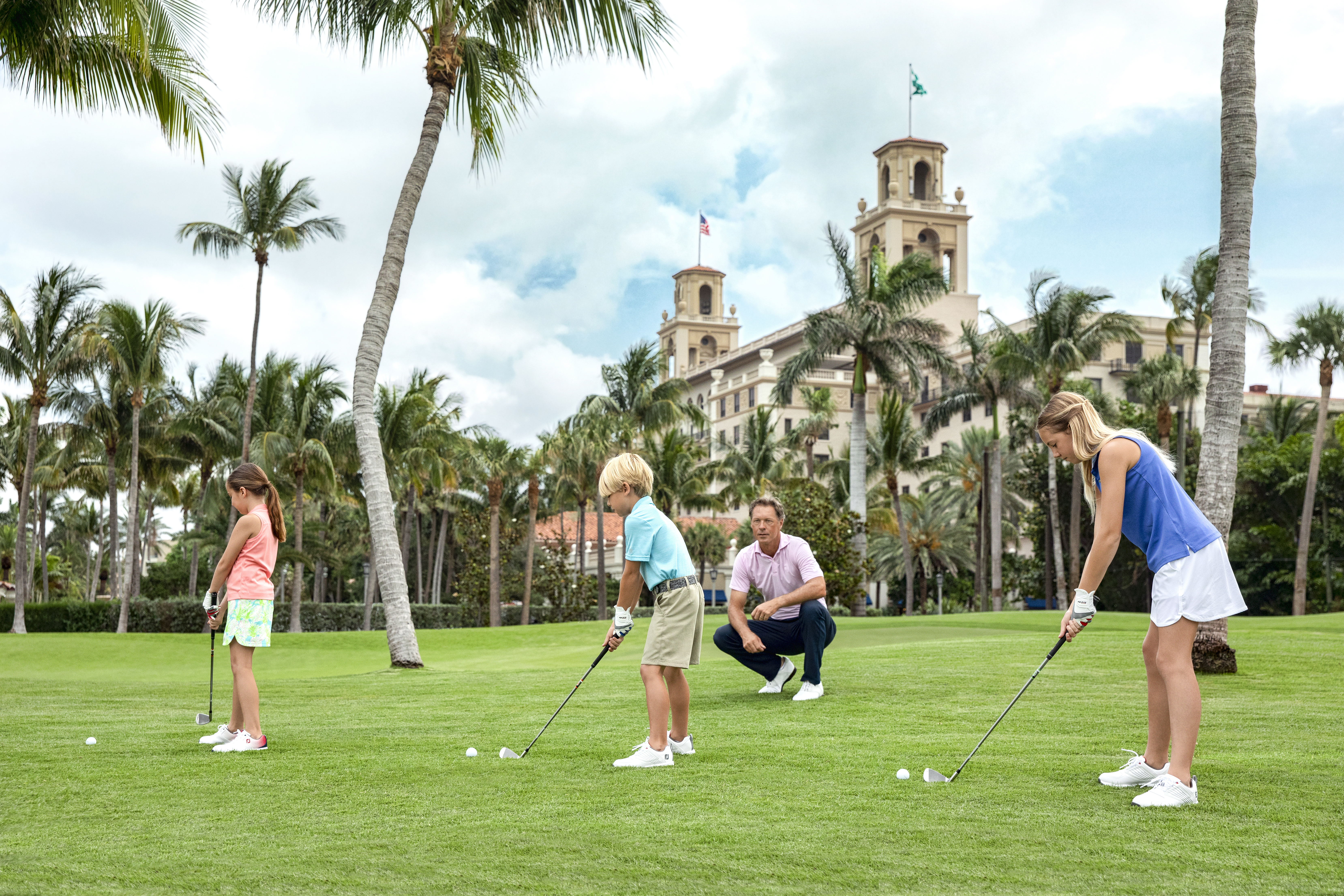 Family of four playing golf on green lawn with palm trees and beige resort building in background.