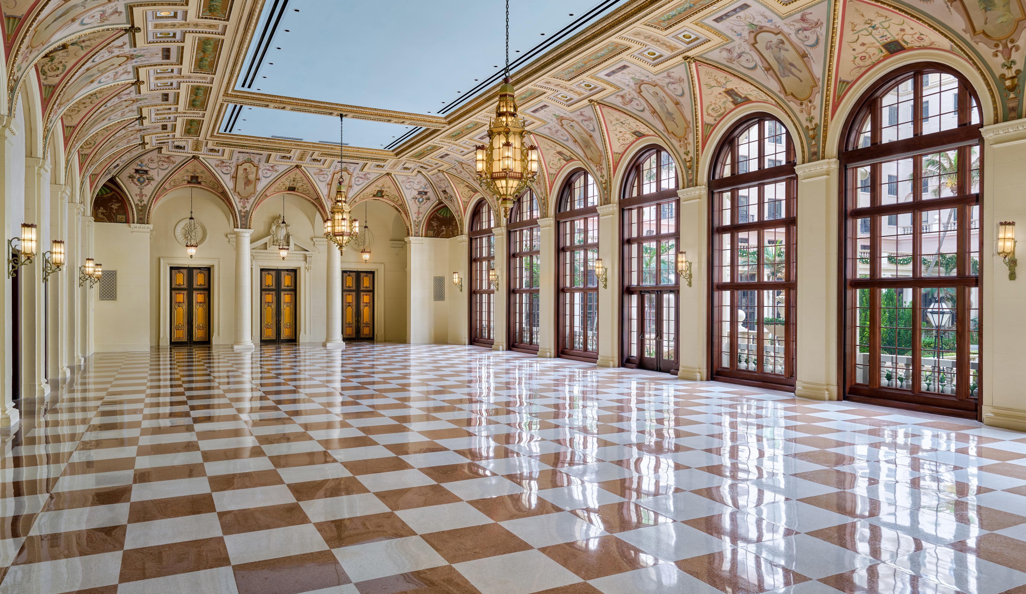 Grand hallway with ornate frescoed ceiling, arched windows, checkered marble floor, and hanging chandeliers.
