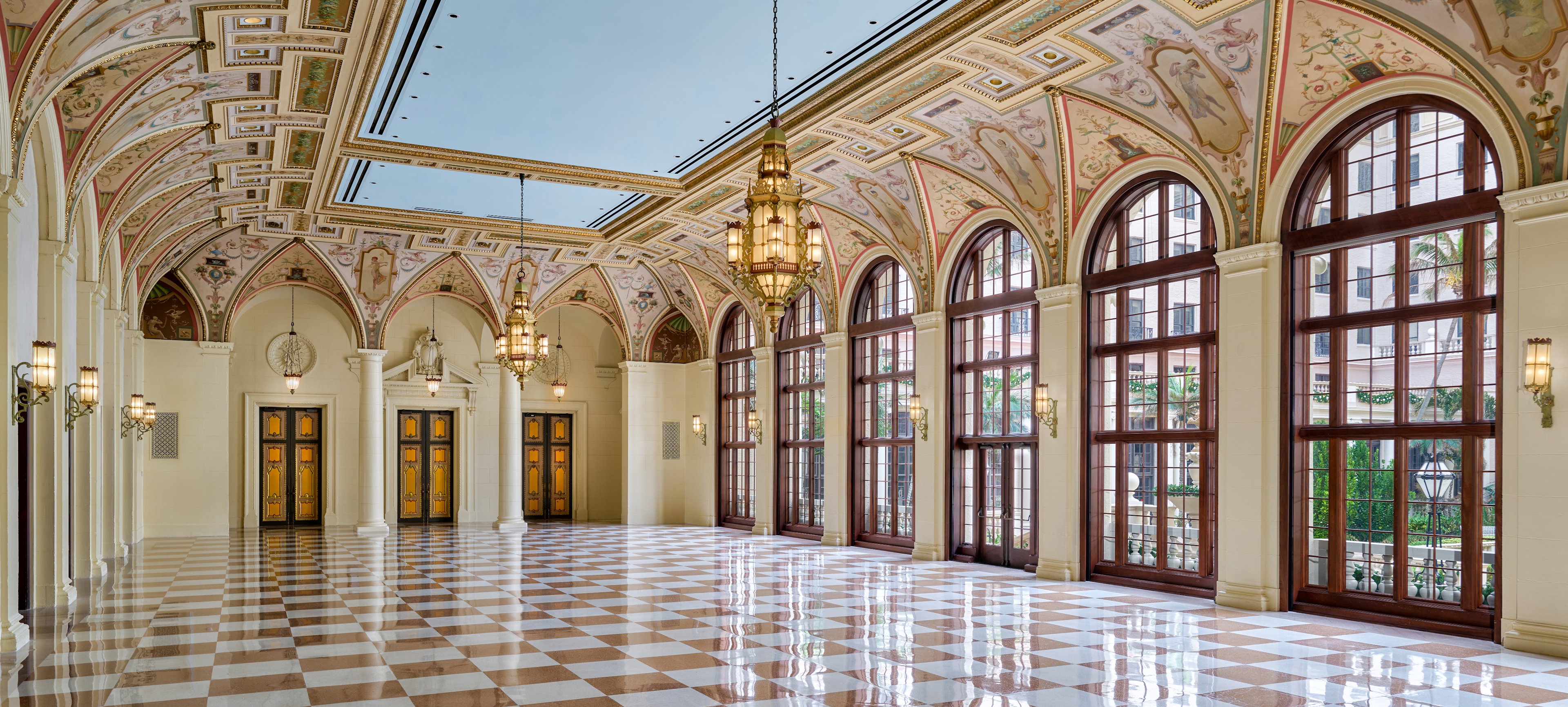 Grand hallway with ornate frescoed ceiling, arched windows, checkered marble floor, and hanging chandeliers.