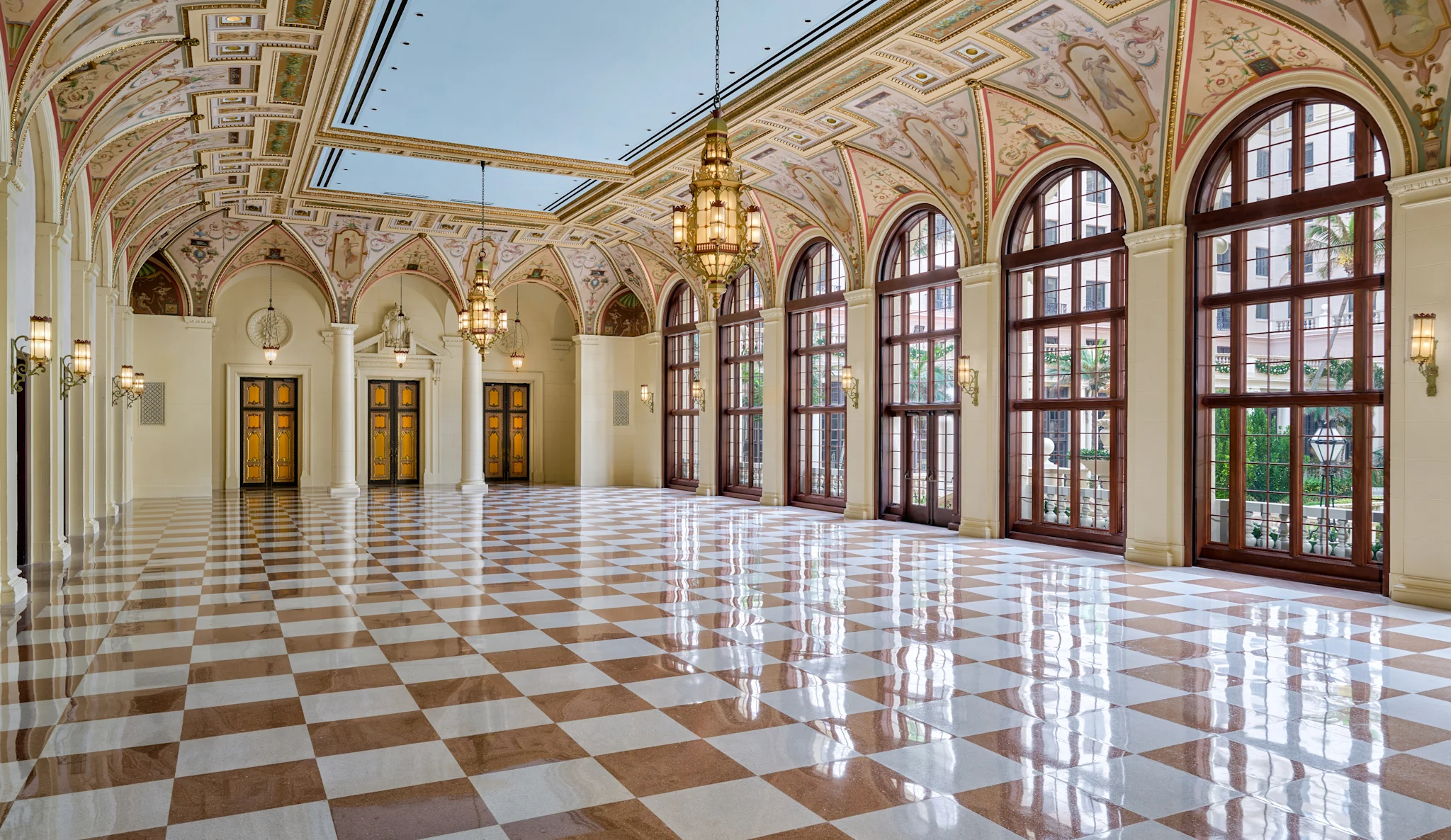 Grand hallway with ornate frescoed ceiling, arched windows, checkered marble floor, and hanging chandeliers.