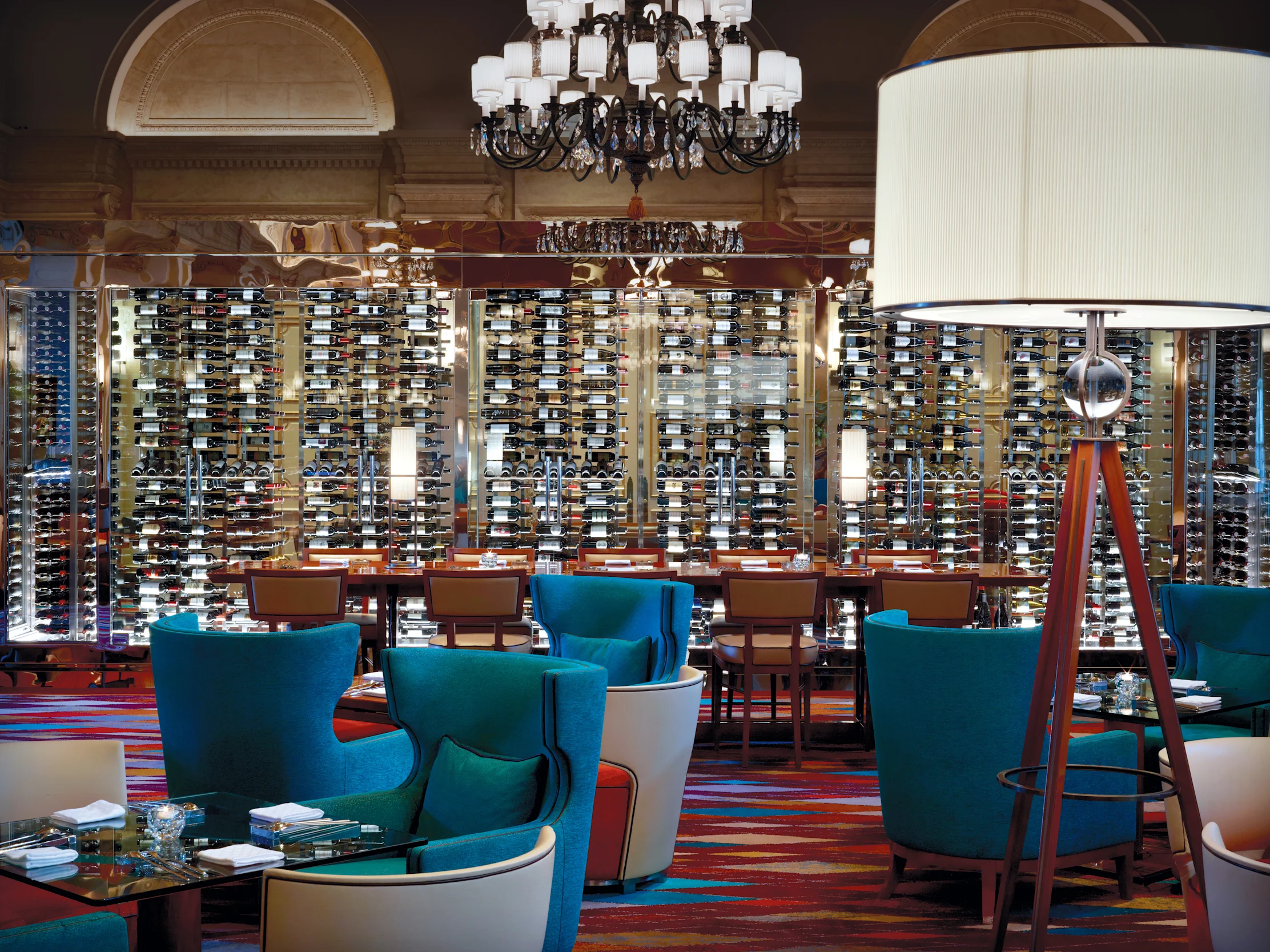 Elegant restaurant interior with turquoise chairs, large white lampshade, chandelier, and illuminated wine cellar wall display.
