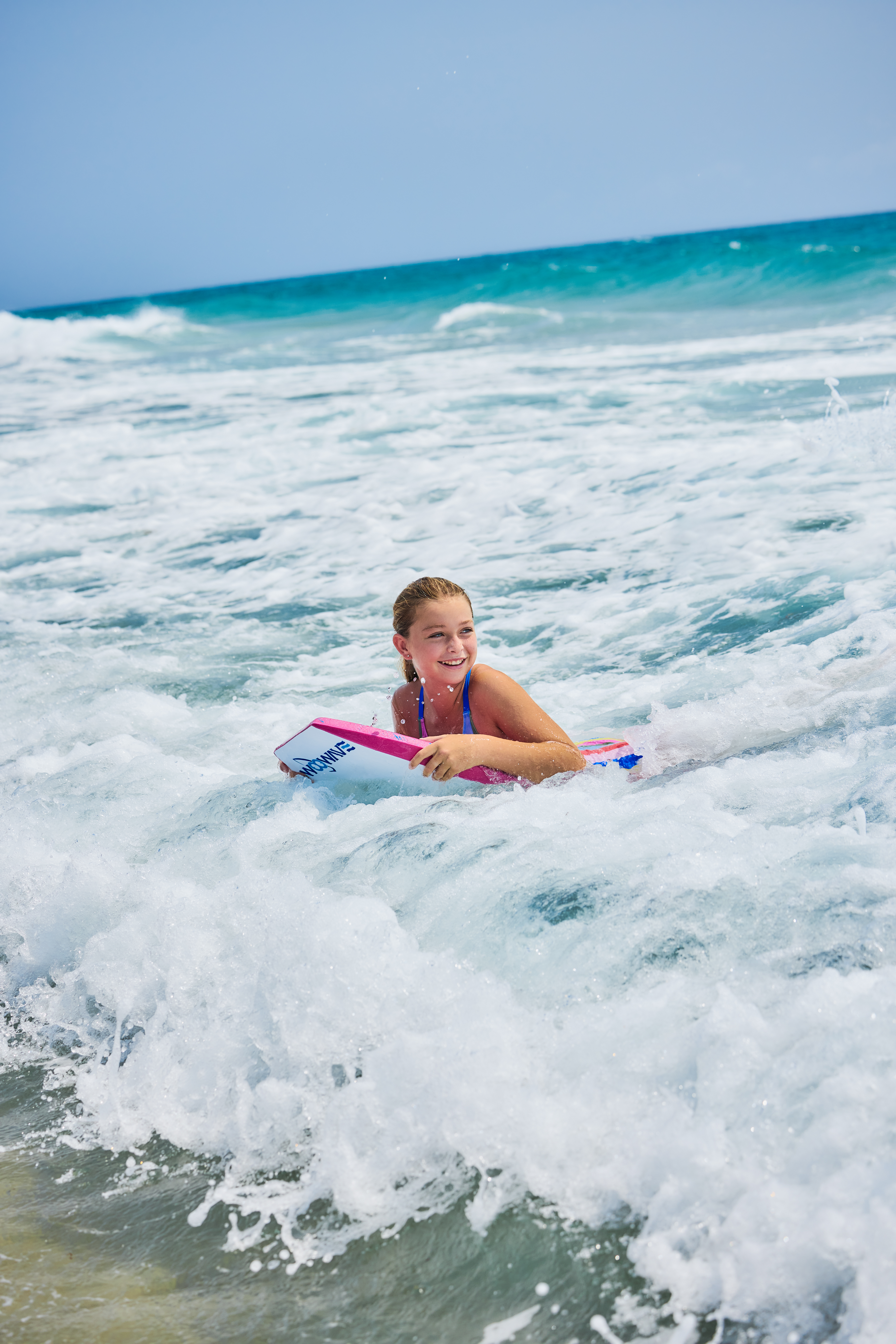 Young girl smiling while bodyboarding in turquoise ocean waves under clear blue sky