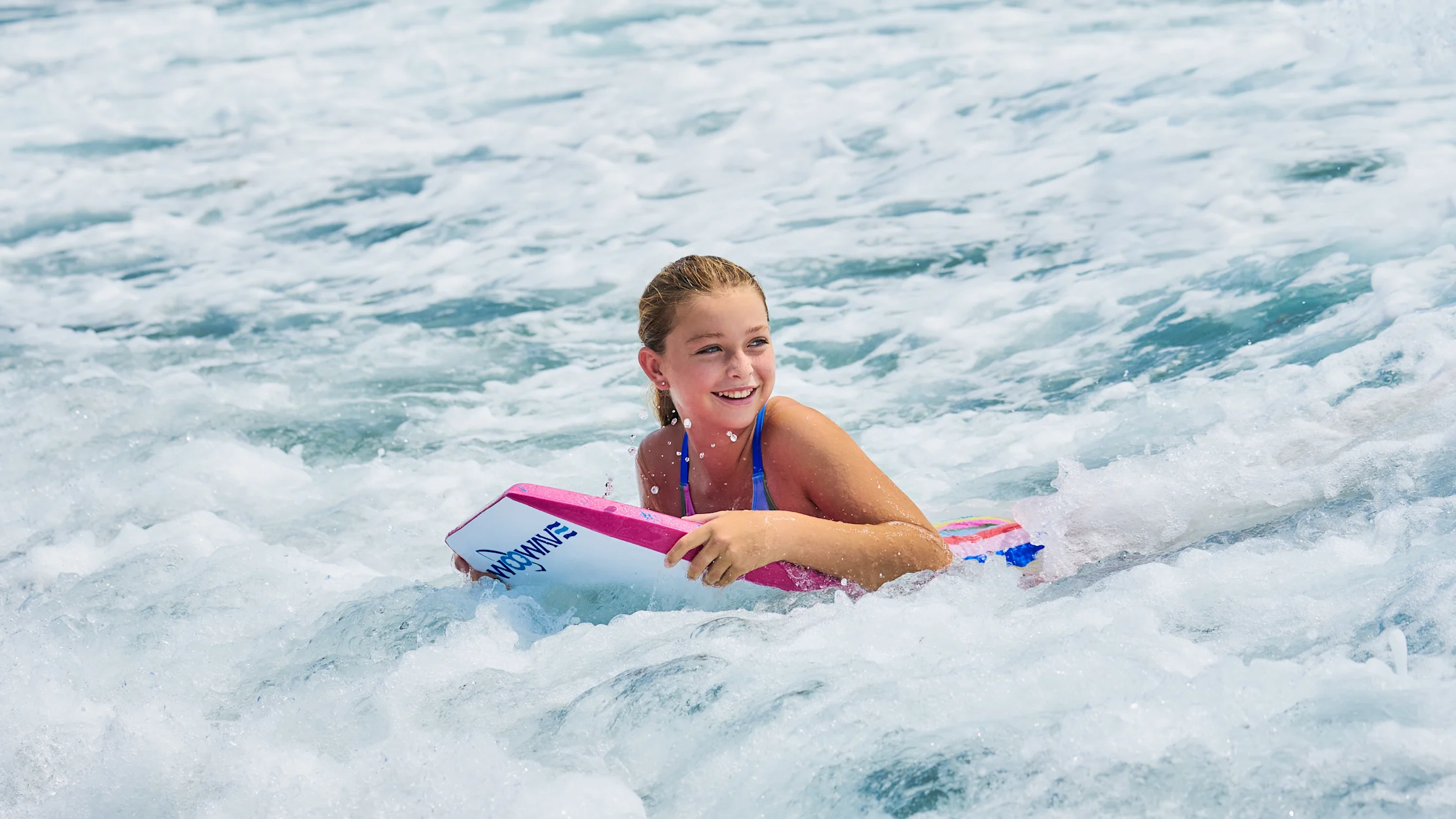 Young girl smiling while bodyboarding in turquoise ocean waves under clear blue sky