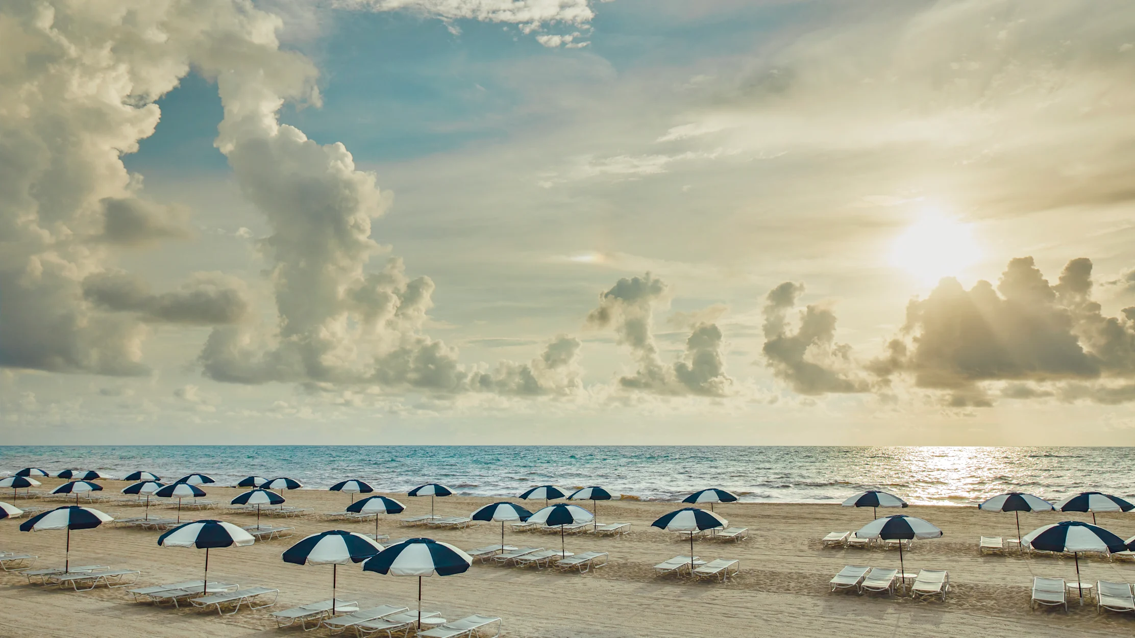 Beach with blue and white striped umbrellas and lounge chairs under a cloudy sky at sunset.