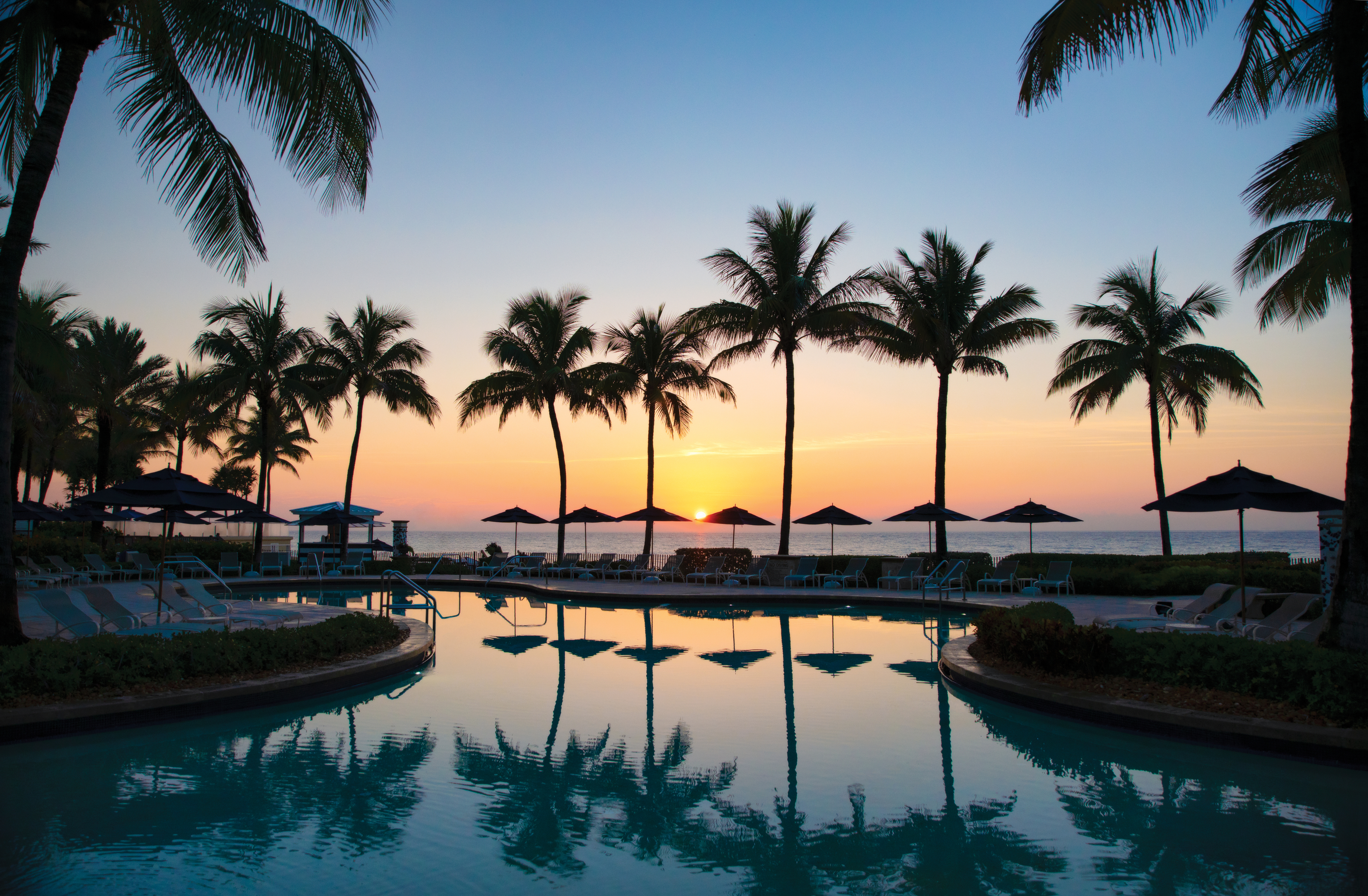Tropical resort pool at sunset with palm trees silhouetted against orange and blue sky.