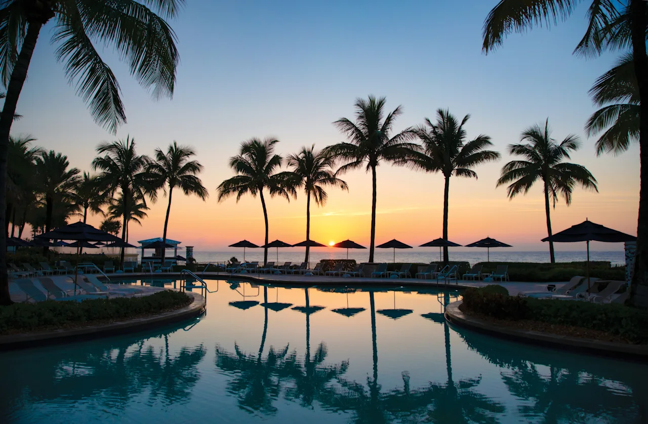 Tropical resort pool at sunset with palm trees silhouetted against orange and blue sky.