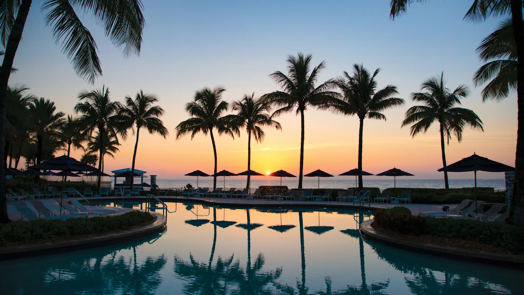 Tropical resort pool at sunset with palm trees silhouetted against orange and blue sky.