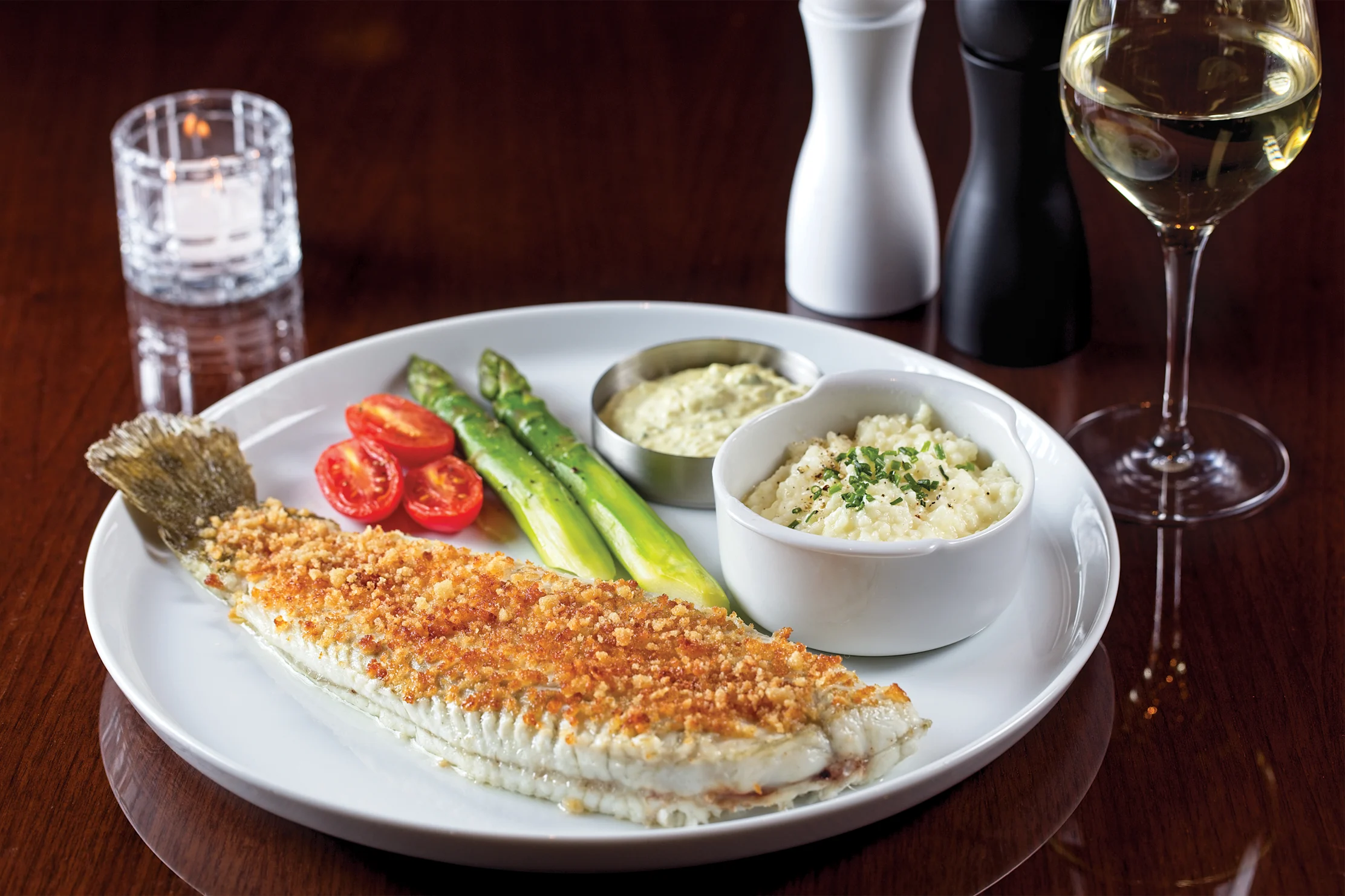 Breaded fish fillet with asparagus, tomatoes, and side dishes on white plate with white wine glass.