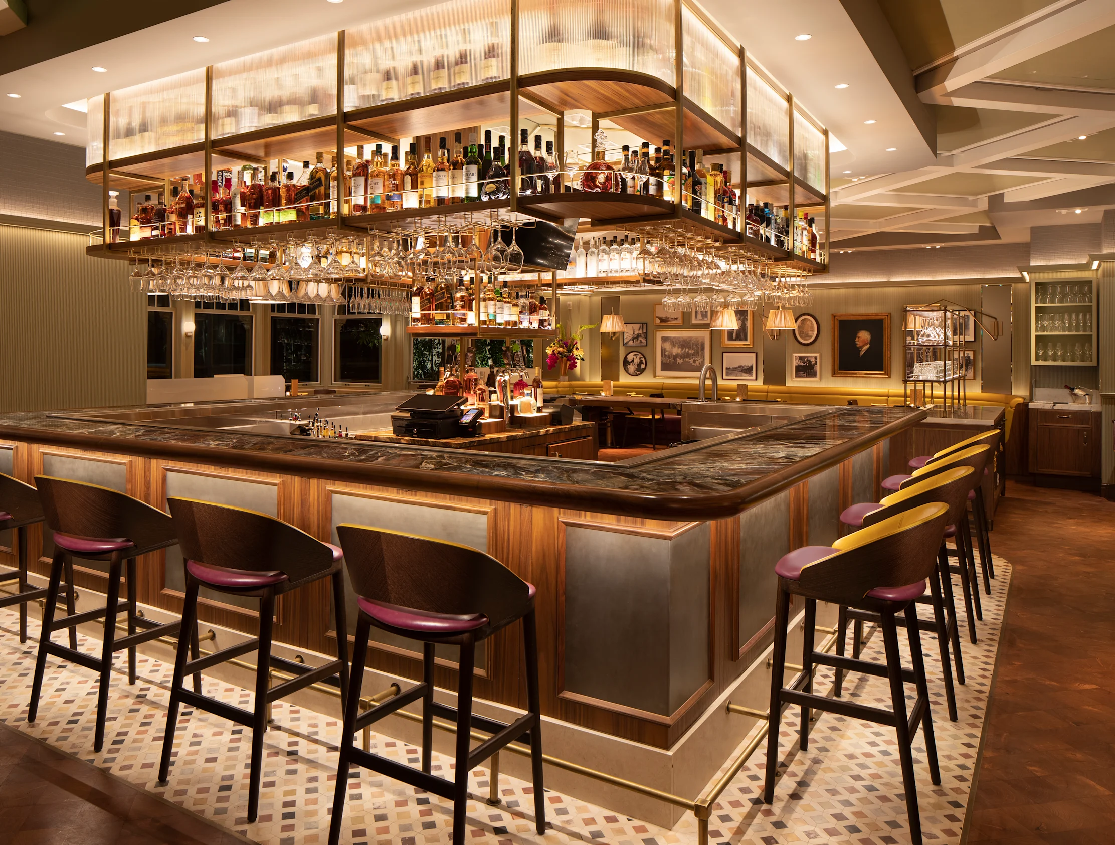 Upscale bar with overhead liquor display, hanging glassware, marble countertop, and purple-cushioned stools.
