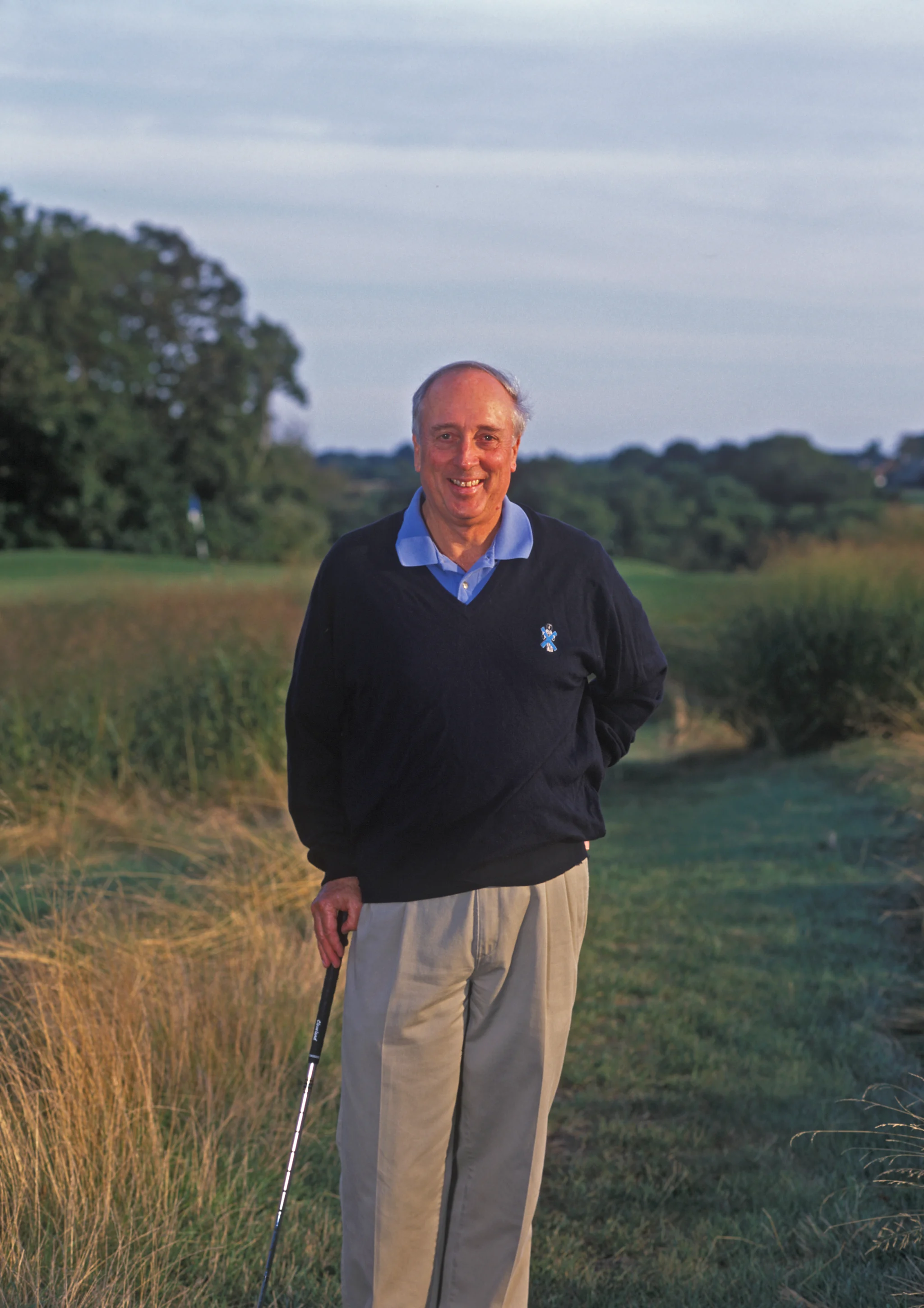 Smiling man in navy sweater and khaki pants holding golf club on course with green grass and trees.