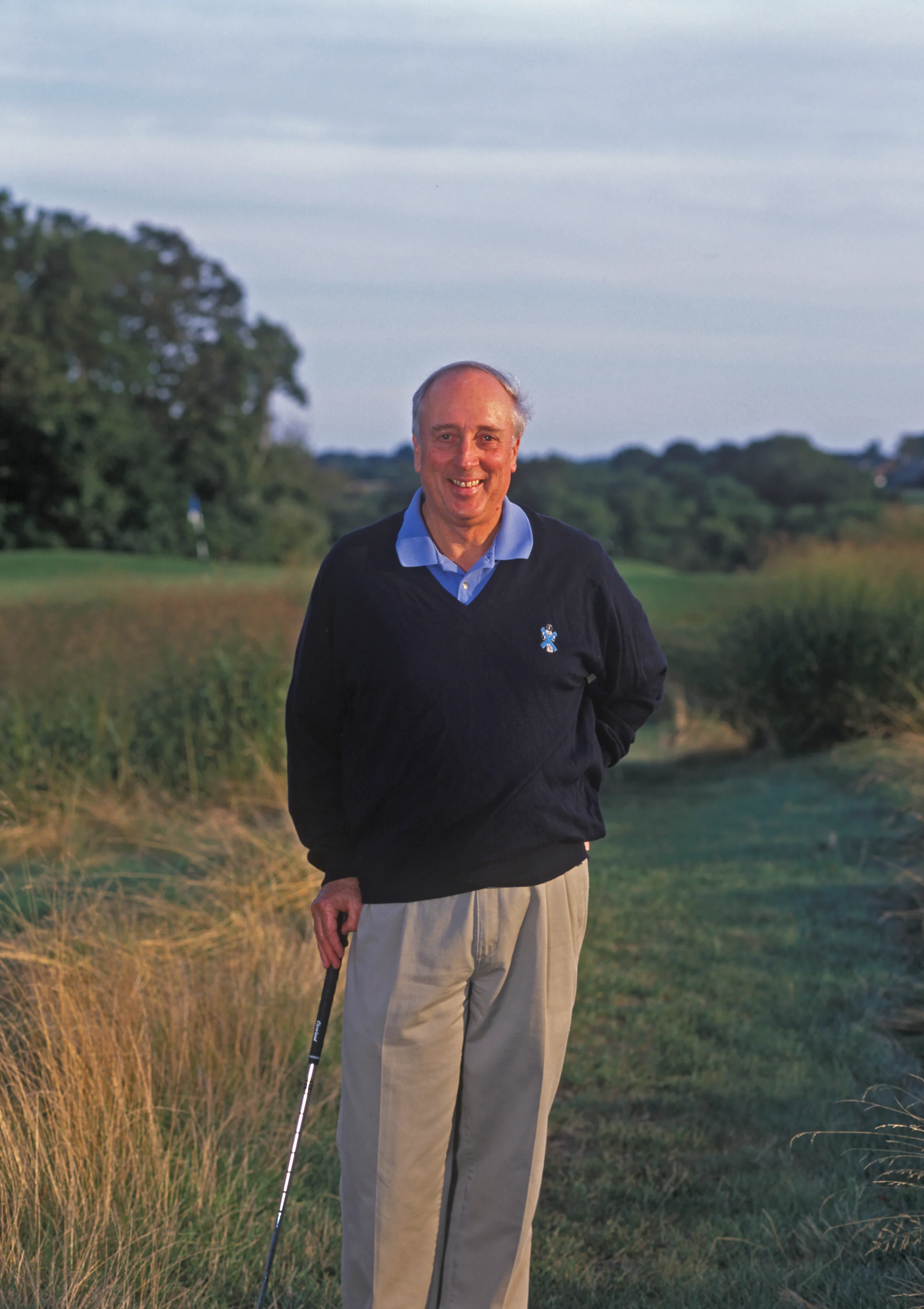 Smiling man in navy sweater and khaki pants holding golf club on course with green grass and trees.