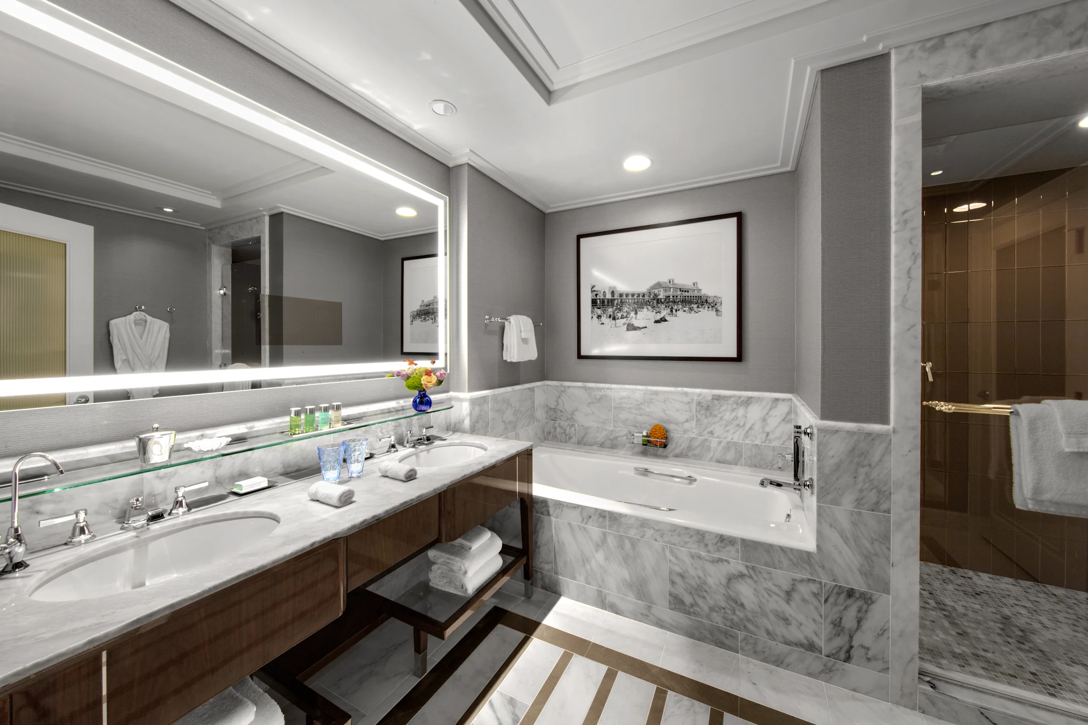 Luxury hotel bathroom with marble vanity, backlit mirror, soaking tub, and gray walls with artwork.