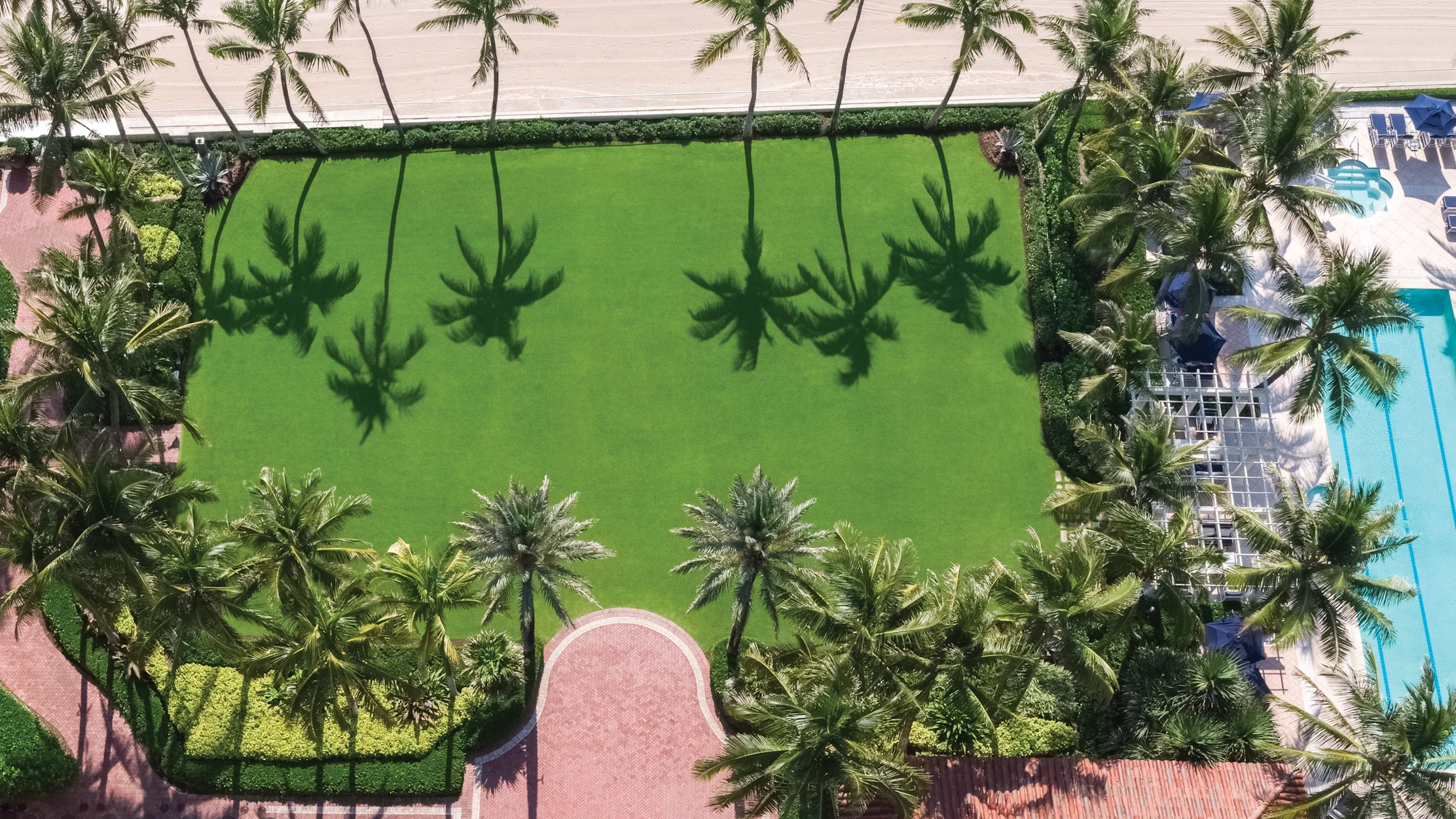 Aerial view of green lawn surrounded by palm trees with beach and pool visible at edges