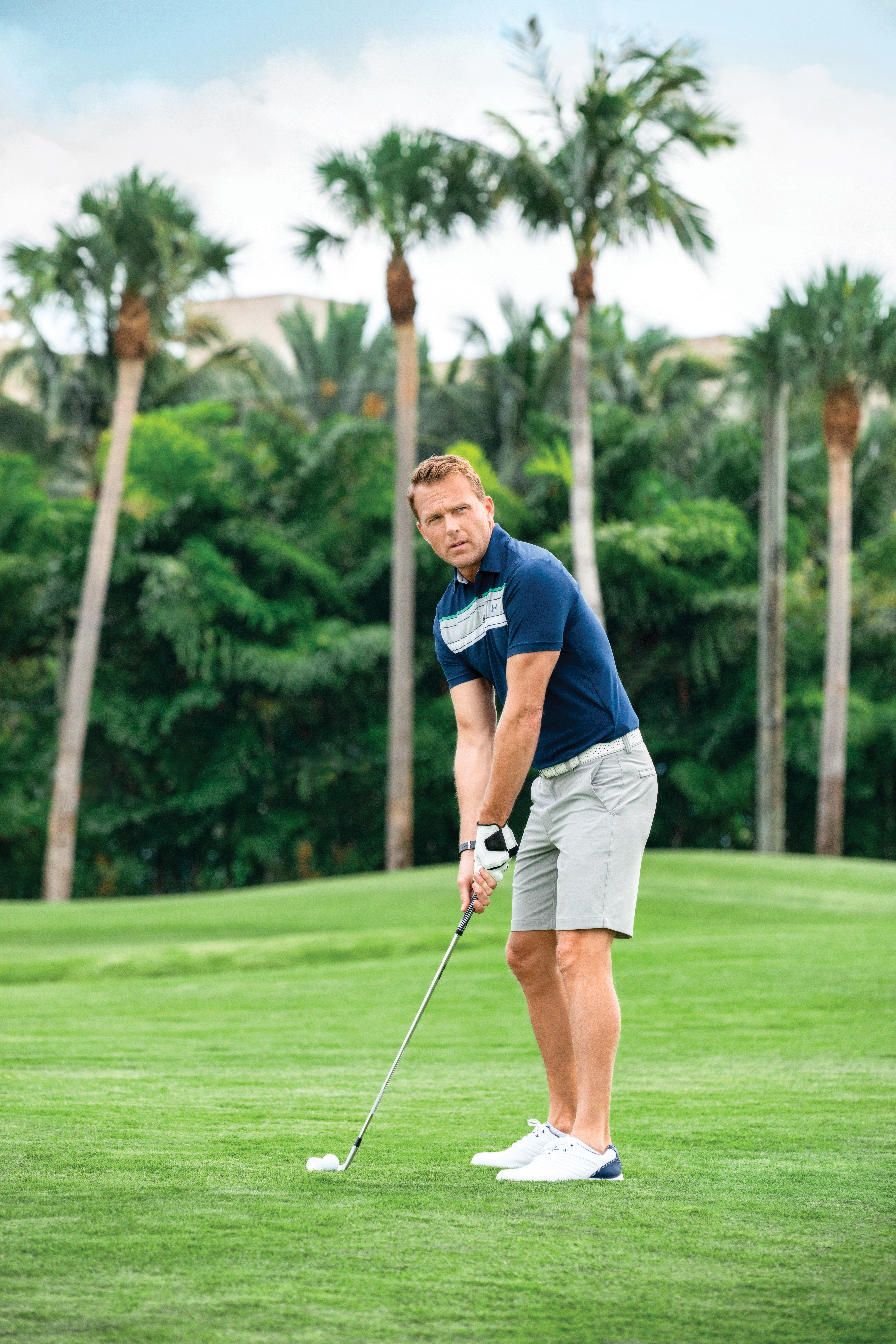 Man in navy polo and gray shorts putting on tropical golf course with palm trees in background.