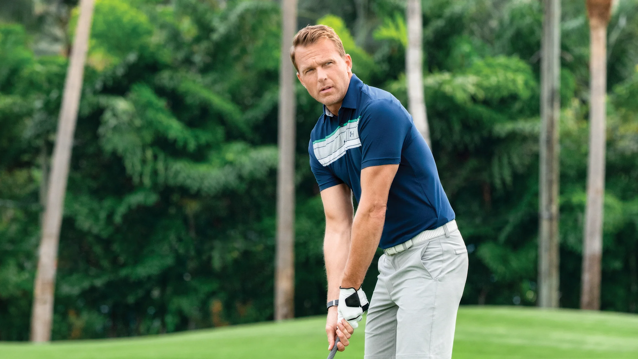 Man in navy polo and gray shorts putting on tropical golf course with palm trees in background.