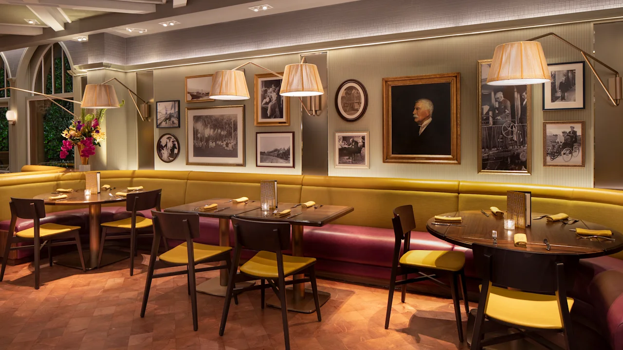 Elegant restaurant interior with yellow banquette seating, vintage portraits on gray walls, and arc lamps.