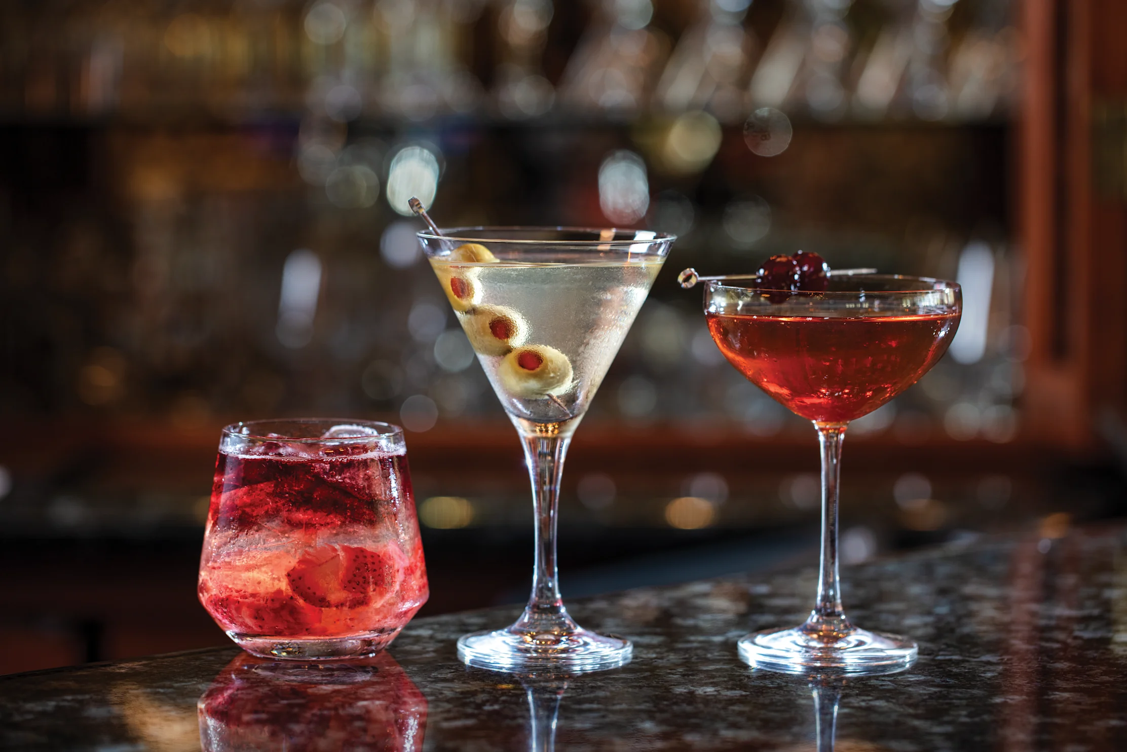 Three cocktails on a bar: a red drink with ice, a martini with olives, and a red coupe with cherry.
