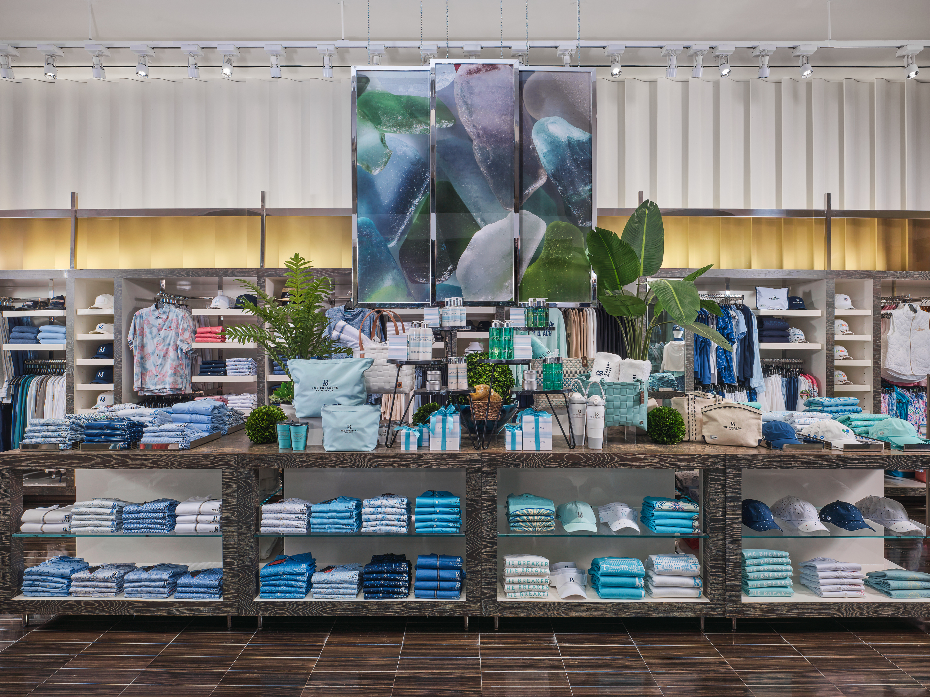 Modern retail store interior with folded clothing in blue and white tones, plants, and sea glass artwork.