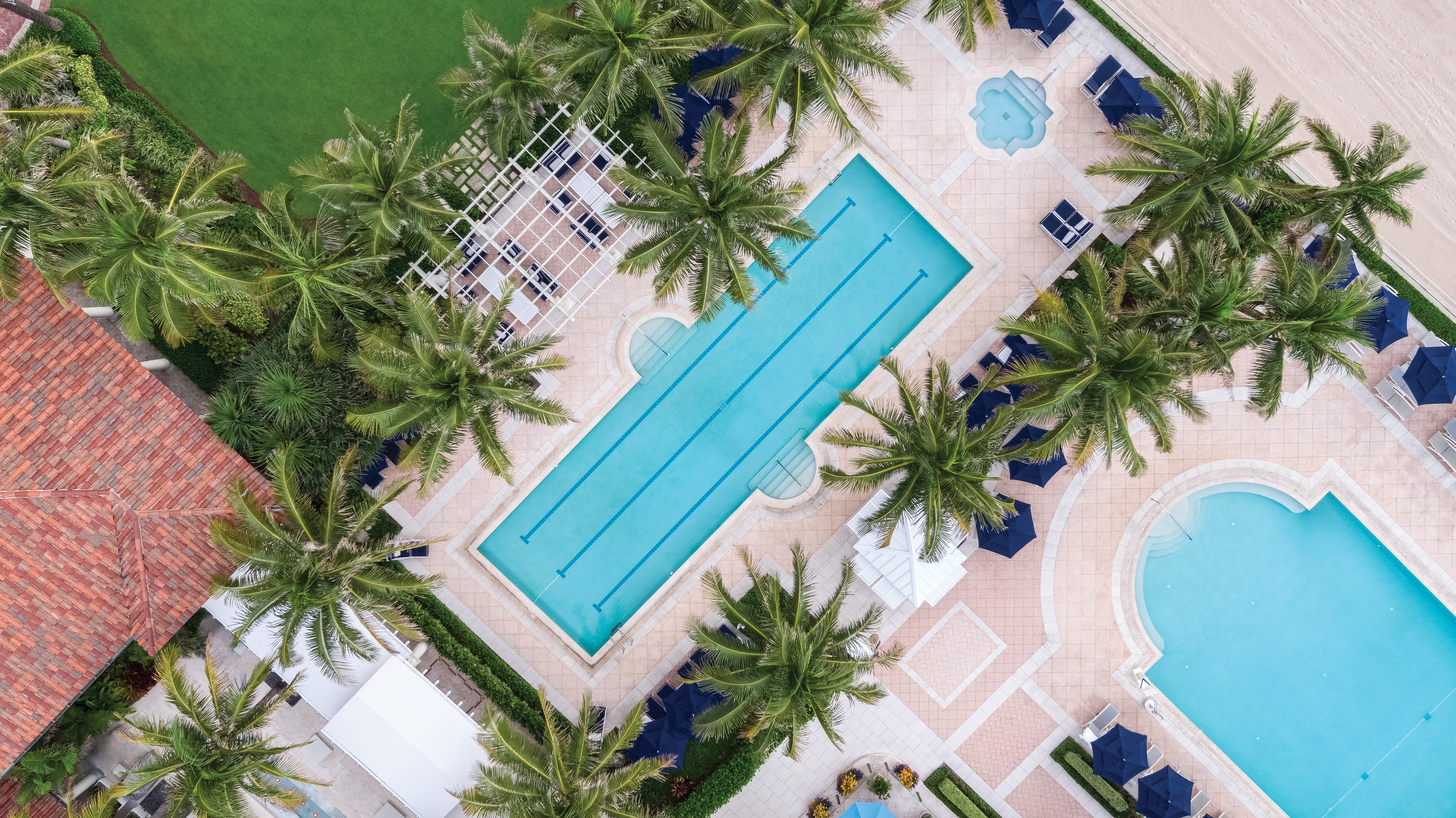 Aerial view of resort pools surrounded by palm trees, blue umbrellas, and beige paving with green lawn.