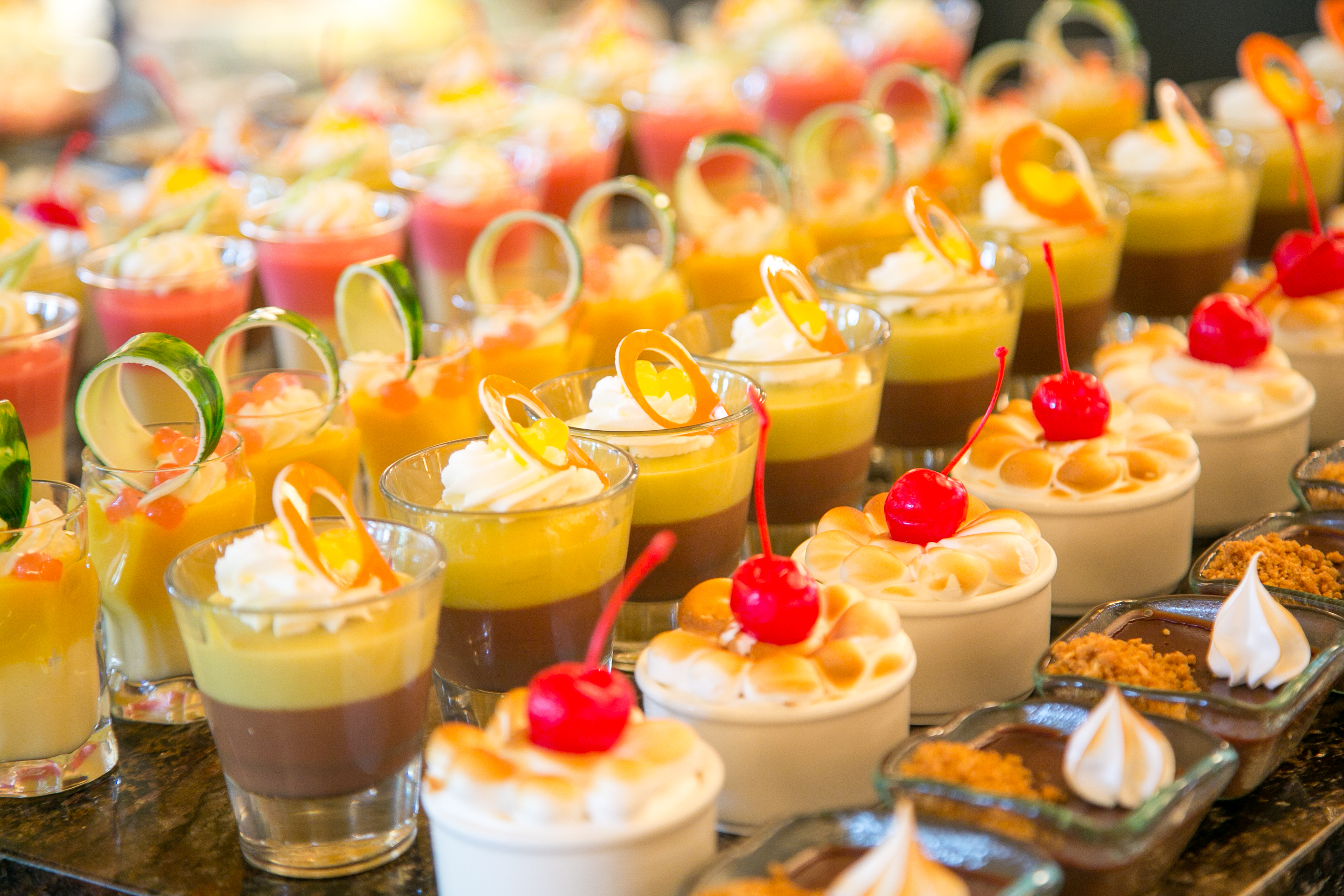 Assorted colorful desserts in small glasses with whipped cream, cherries, and citrus garnishes.