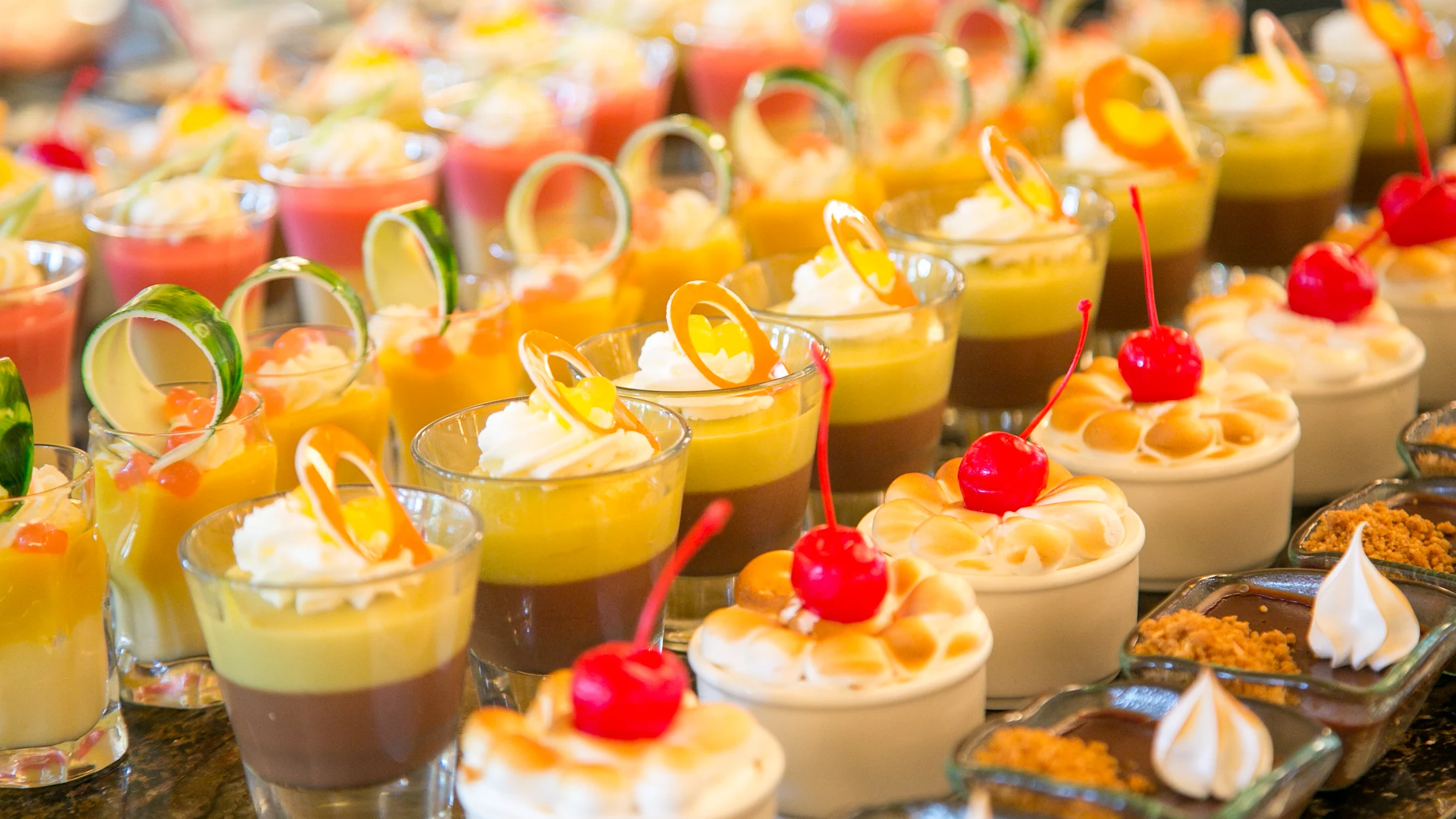 Assorted colorful desserts in small glasses with whipped cream, cherries, and citrus garnishes.