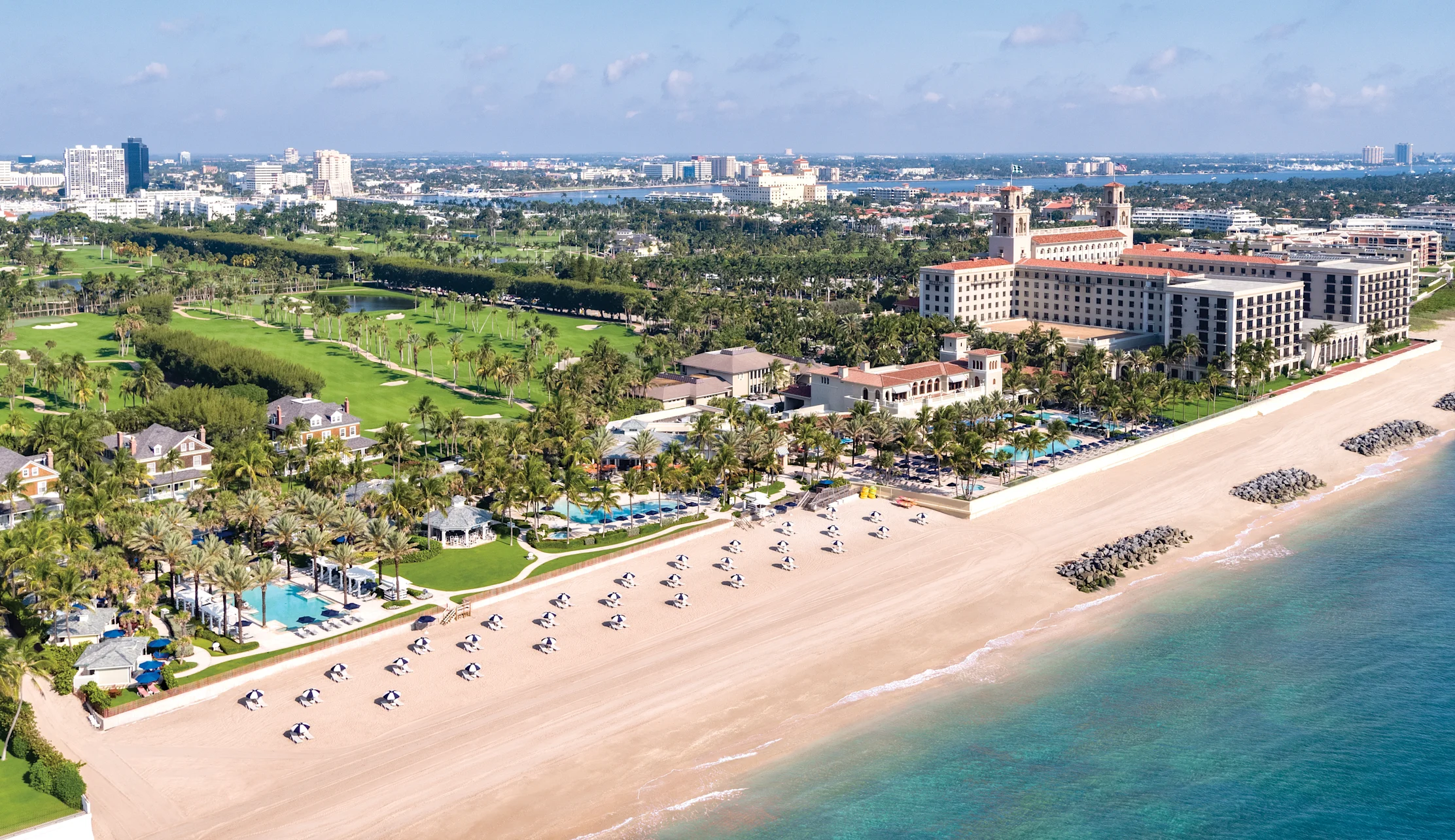 Aerial view of beachfront resort with white sand, turquoise water, palm trees, and golf course.