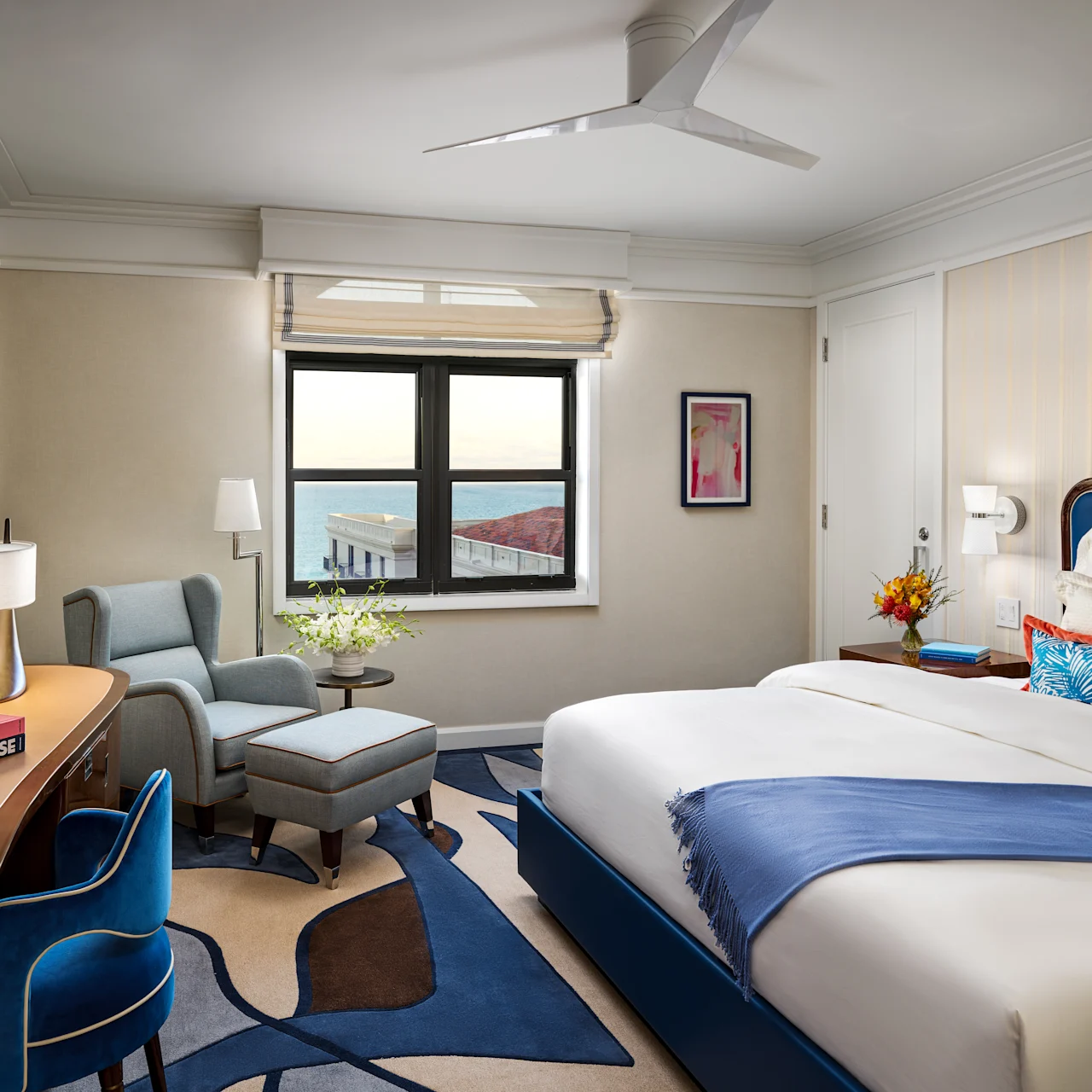Modern hotel room with blue and white decor, featuring a bed, desk, armchair, and window view.