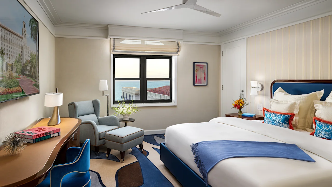 Modern hotel room with blue and white decor, featuring a bed, desk, armchair, and window view.