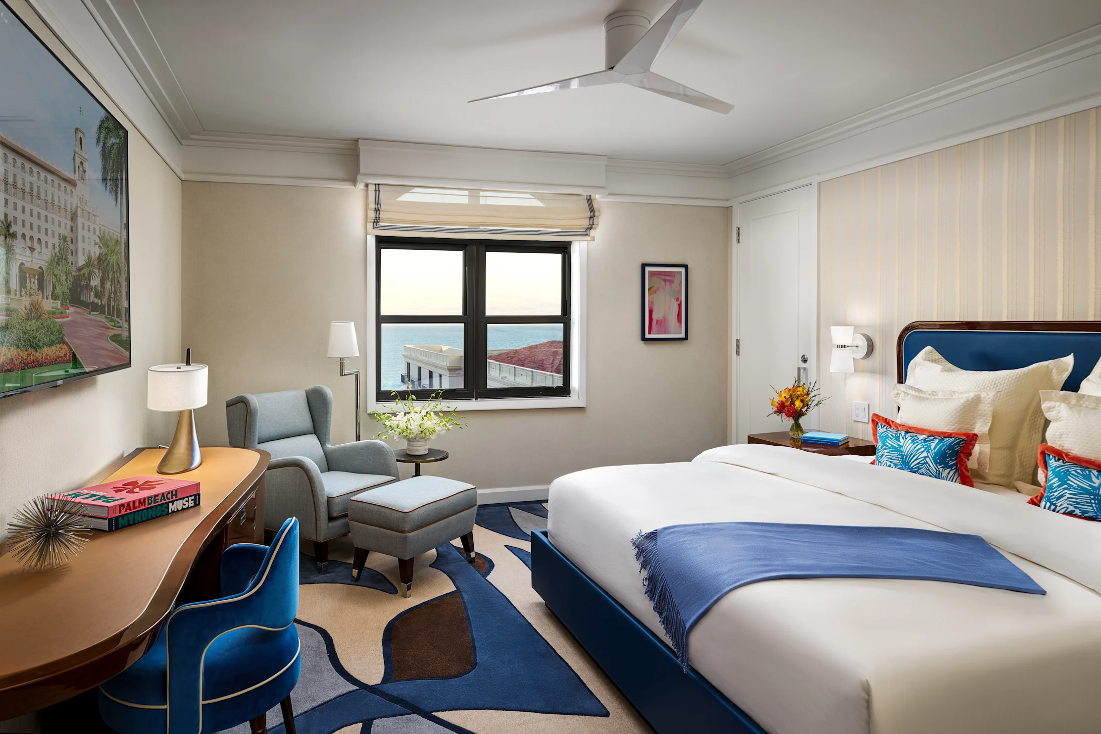 Modern hotel room with blue and white decor, featuring a bed, desk, armchair, and window view.