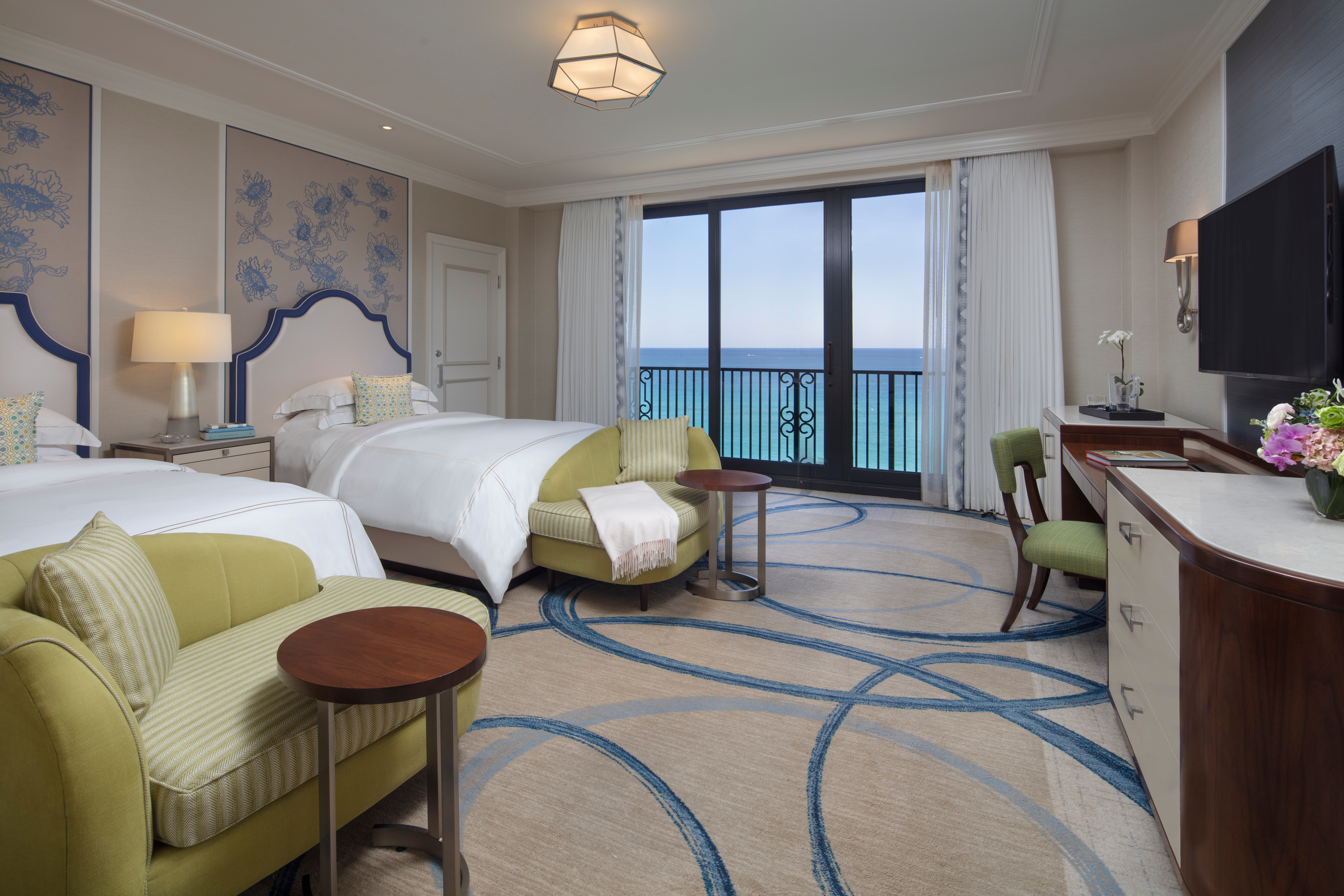 Luxury hotel room with two beds, green armchairs, and ocean view through balcony doors.