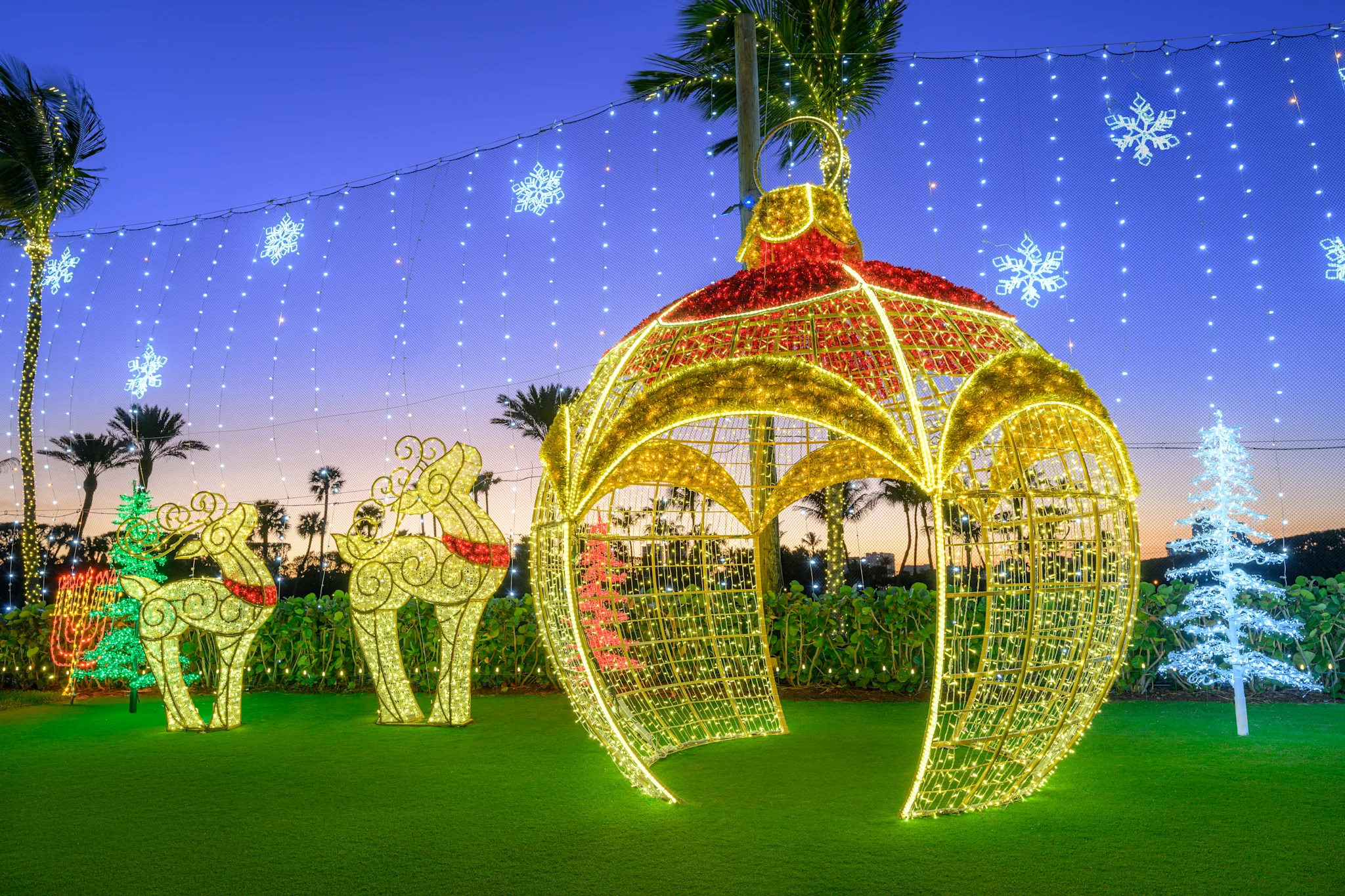 Illuminated Christmas ornament and reindeer decorations on lawn at dusk with palm trees and lights.