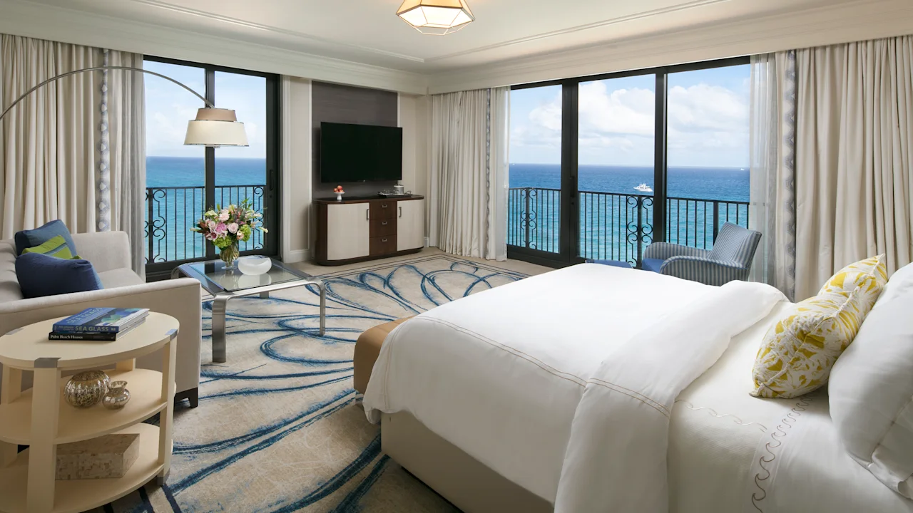 Luxury hotel room with ocean view balcony, white bedding, blue wave-patterned rug, and modern decor.