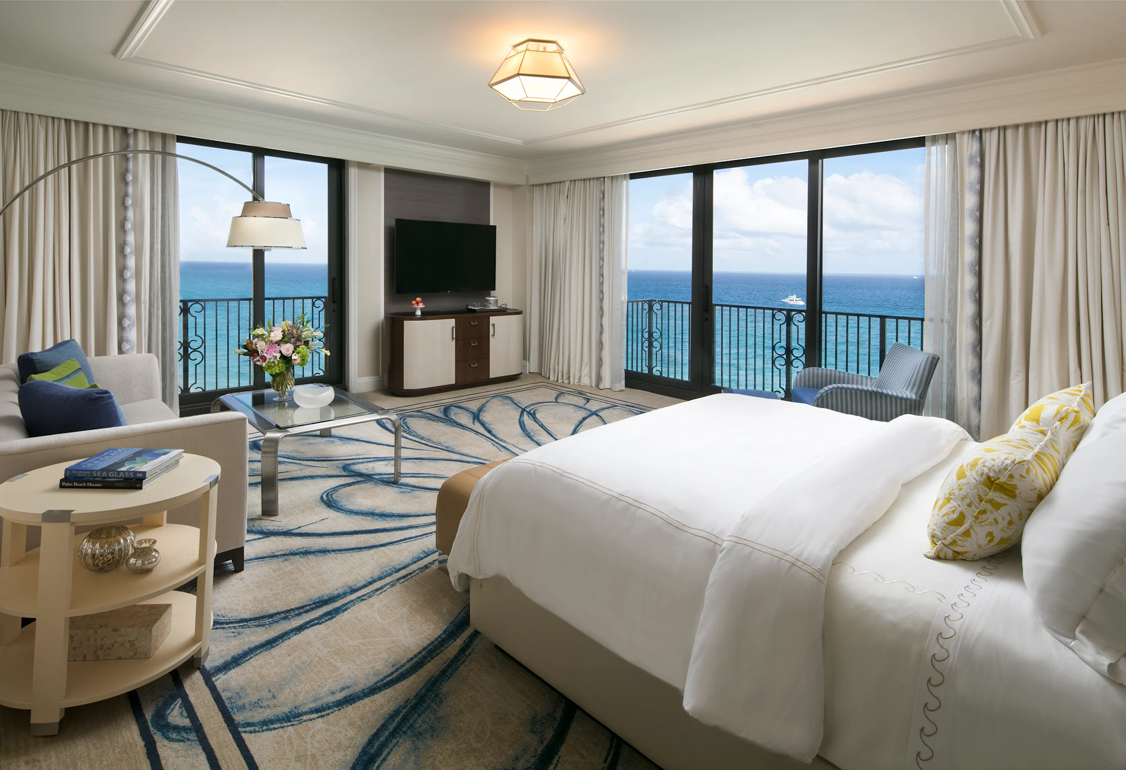 Luxury hotel room with ocean view balcony, white bedding, blue wave-patterned rug, and modern decor.