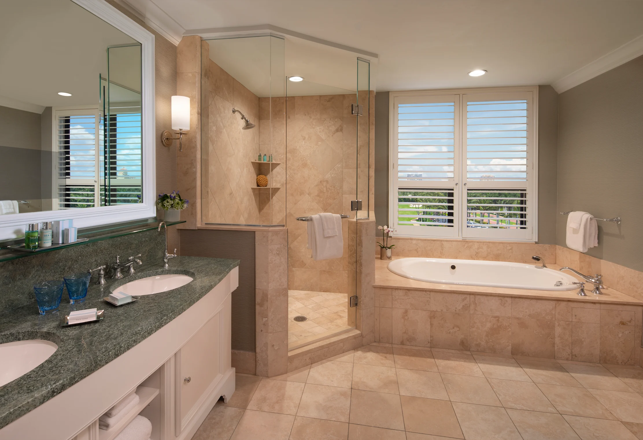 Spacious bathroom with beige tile, glass shower, white soaking tub, dual vanity, and plantation shutters.