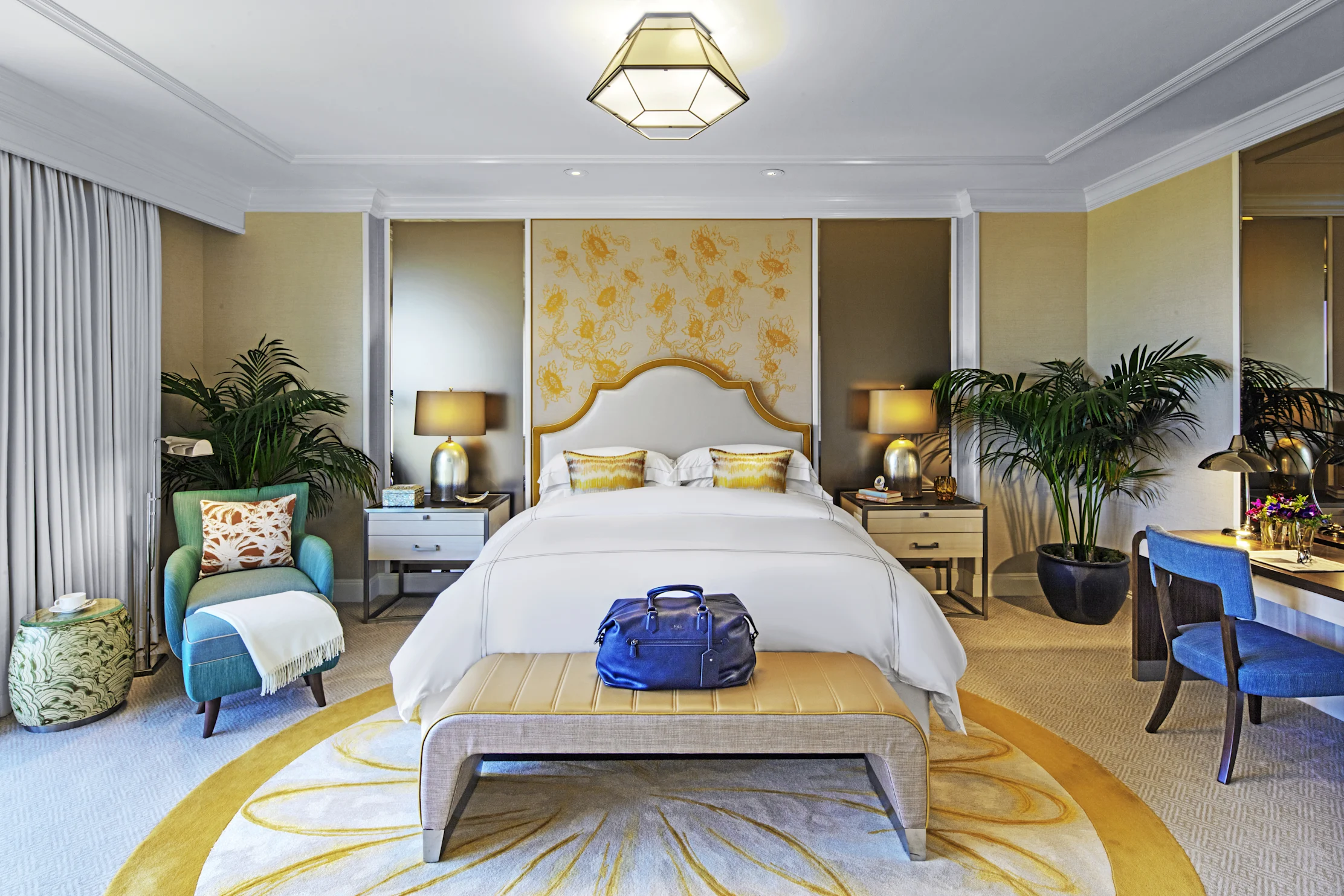 Elegant hotel bedroom with white bed, gold headboard, geometric ceiling light, and palm plants.