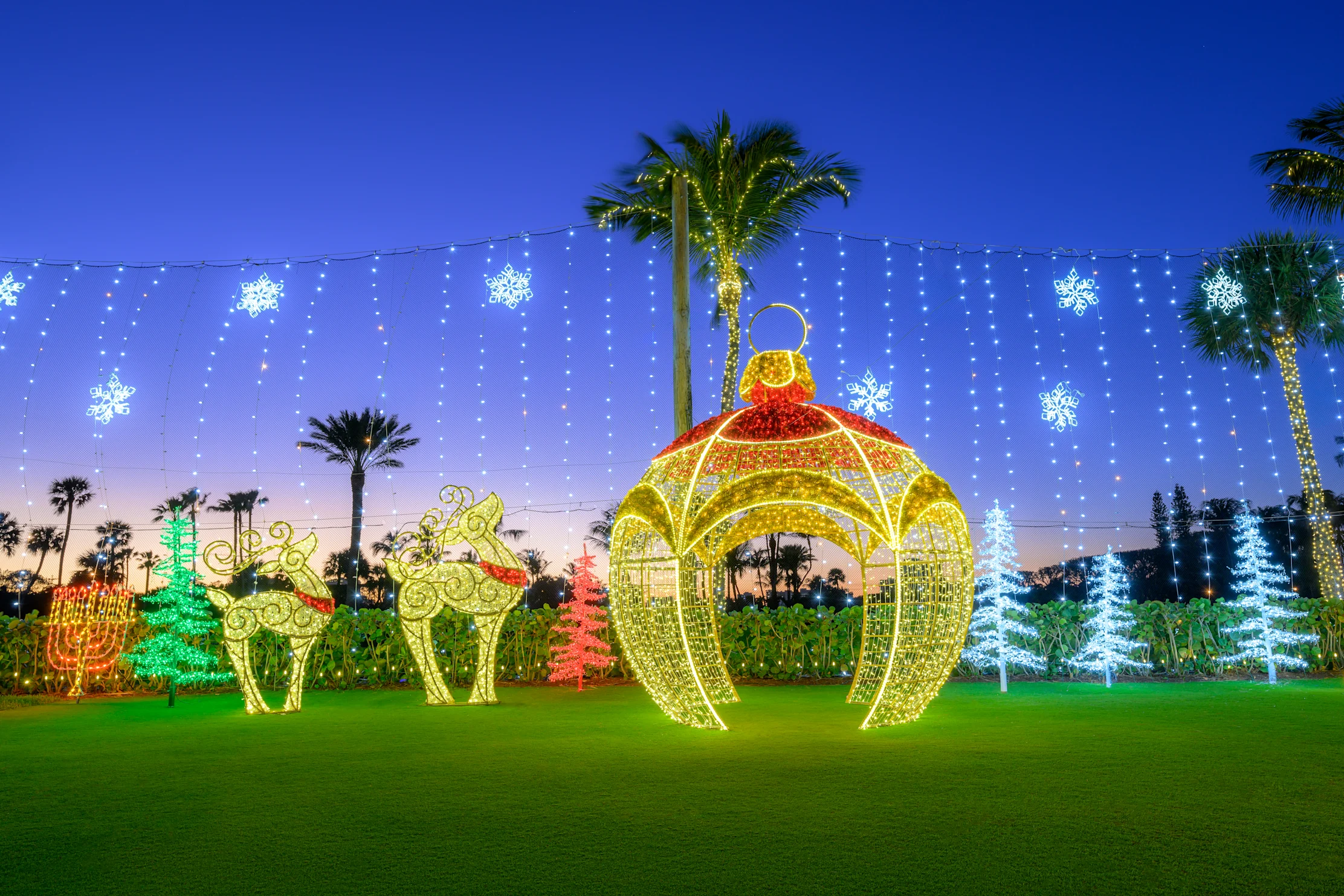 Illuminated Christmas display with giant ornament, reindeer, and snowflakes on lawn with palm trees at dusk.