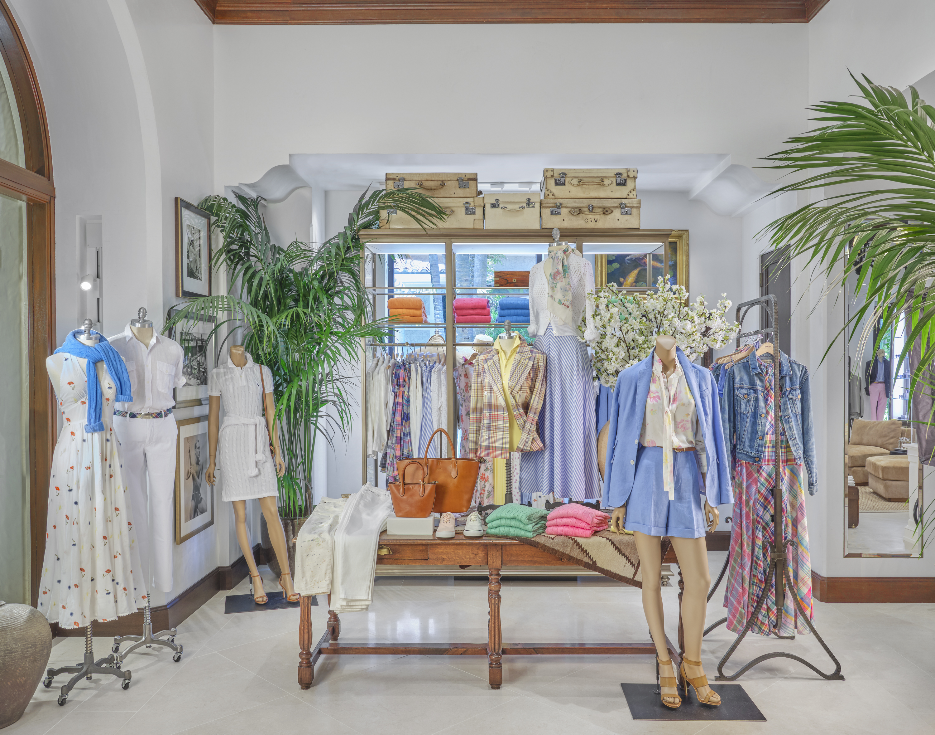 Bright boutique interior with mannequins displaying colorful spring clothing, palm plants, and vintage trunks.