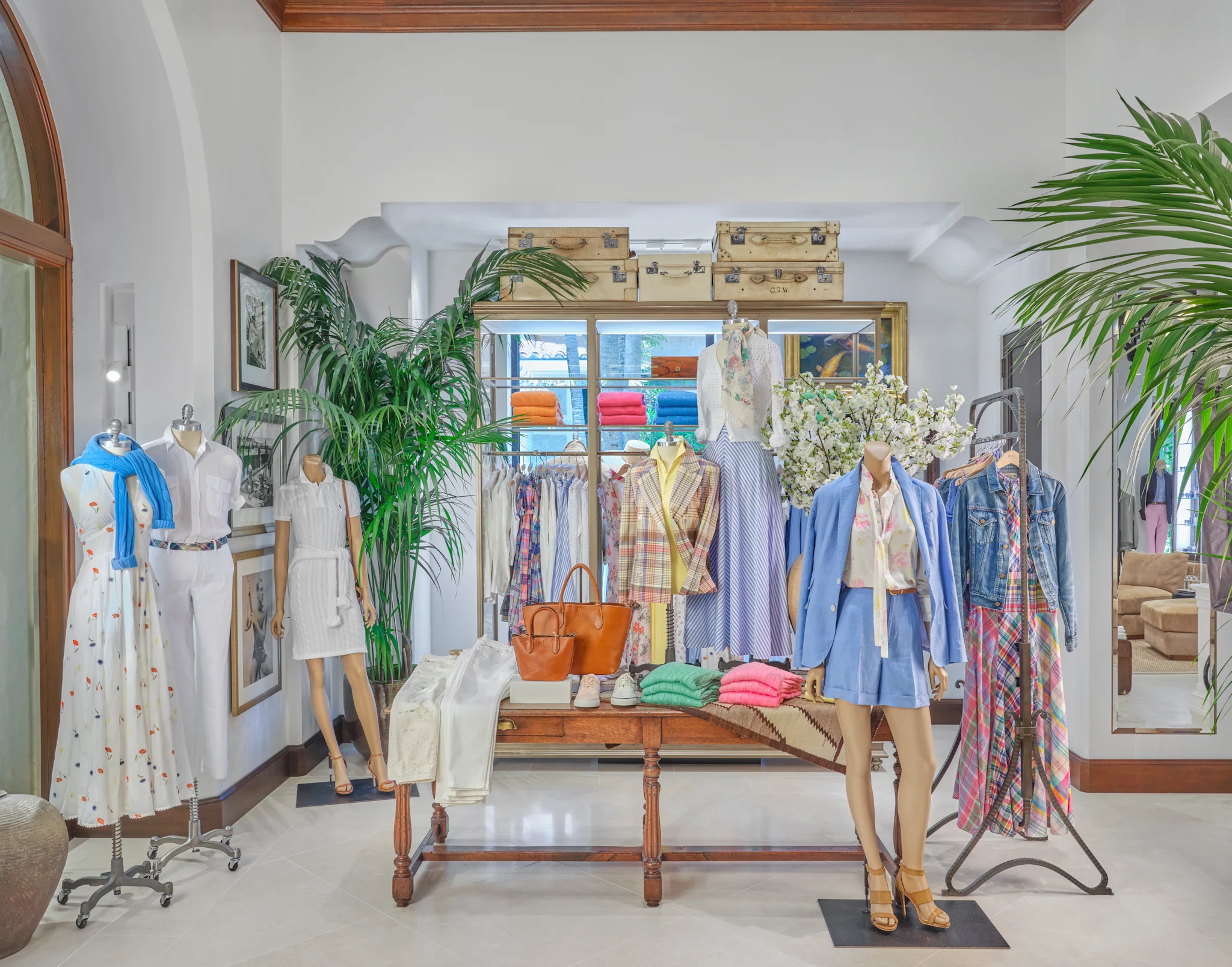 Bright boutique interior with mannequins displaying colorful spring clothing, palm plants, and vintage trunks.
