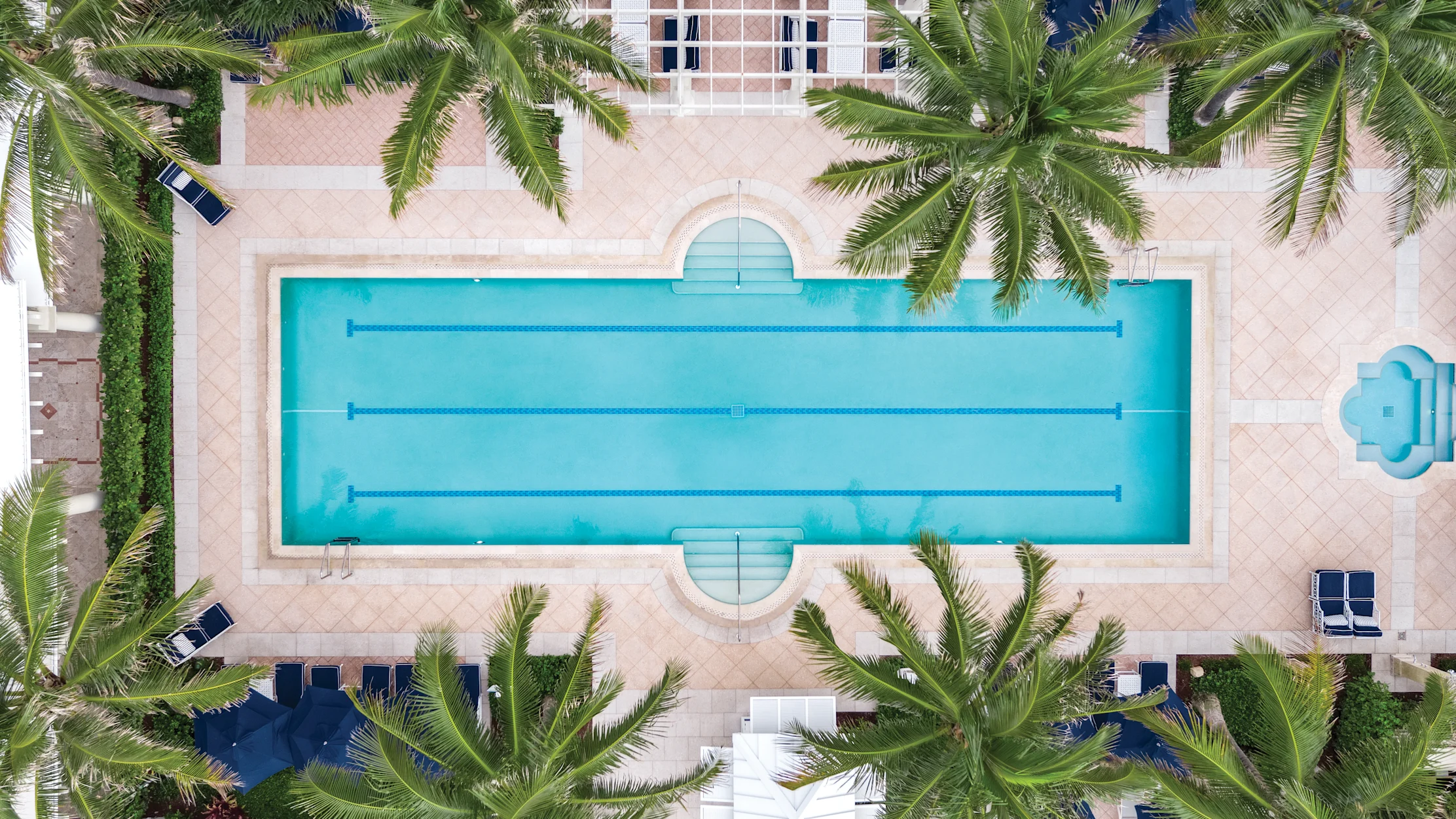 Aerial view of a rectangular swimming pool with lane dividers, surrounded by beige paving and palm trees.