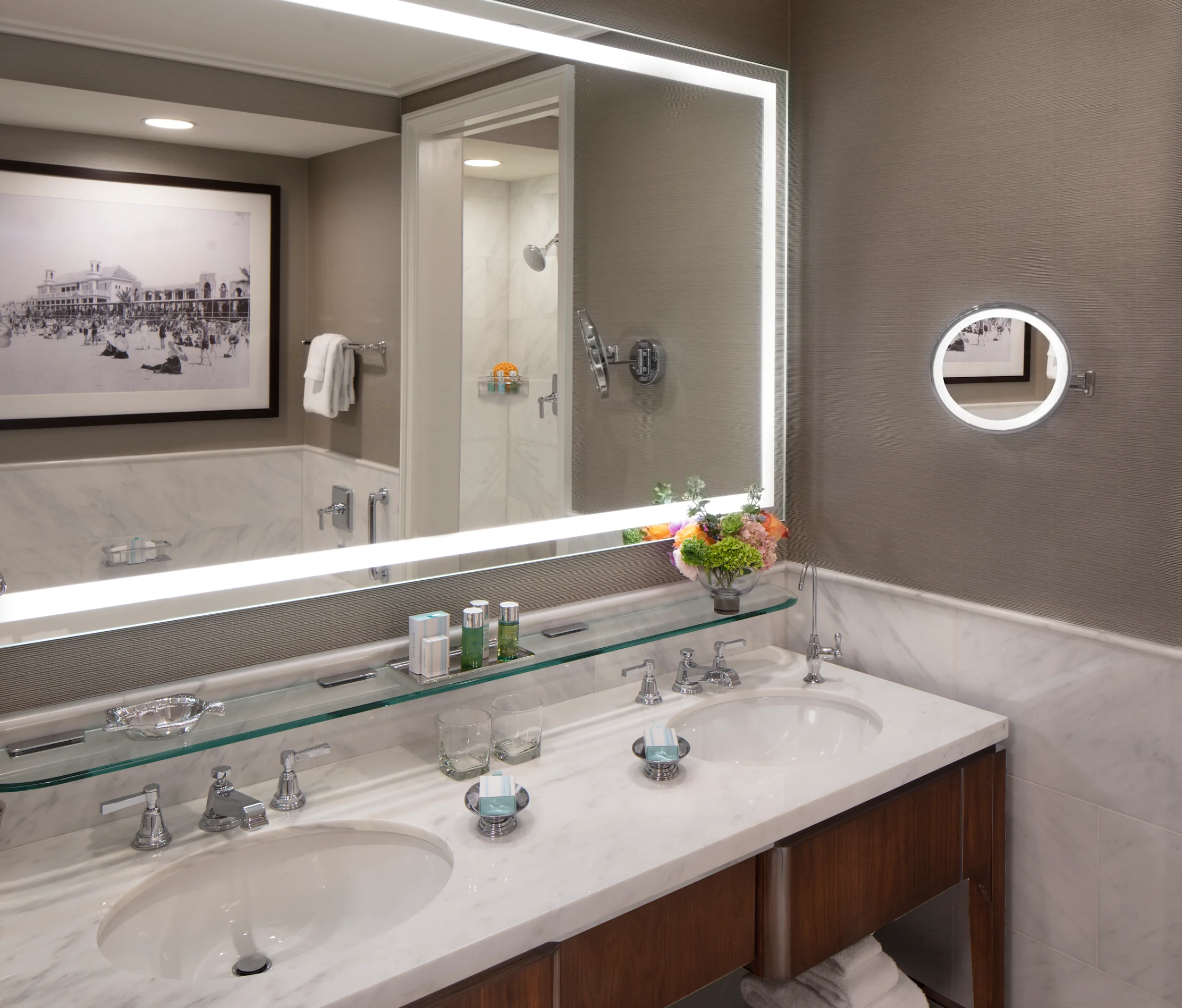 Modern hotel bathroom with double vanity, marble countertops, backlit mirror, and walk-in shower.