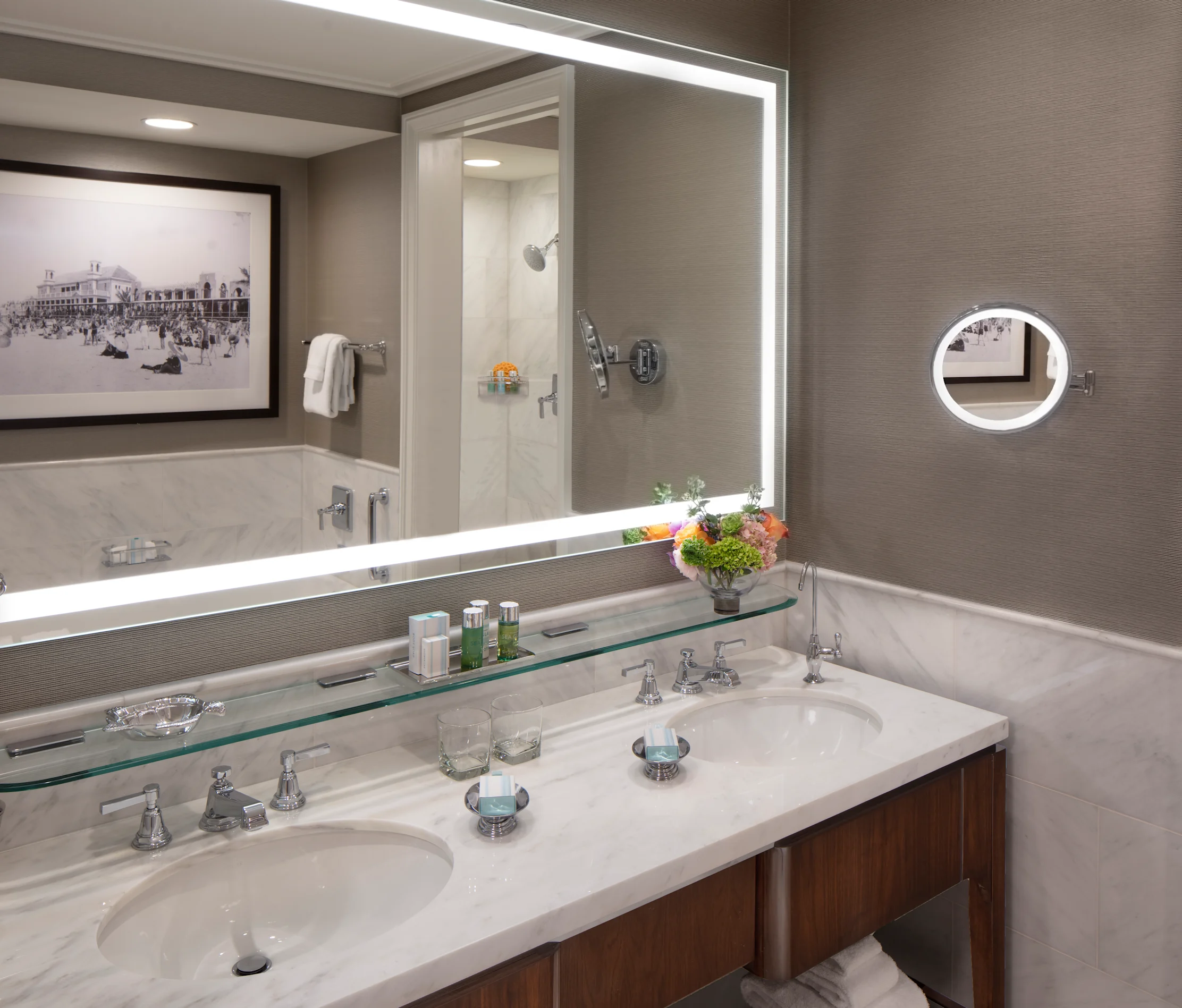Modern hotel bathroom with double vanity, marble countertops, backlit mirror, and walk-in shower.