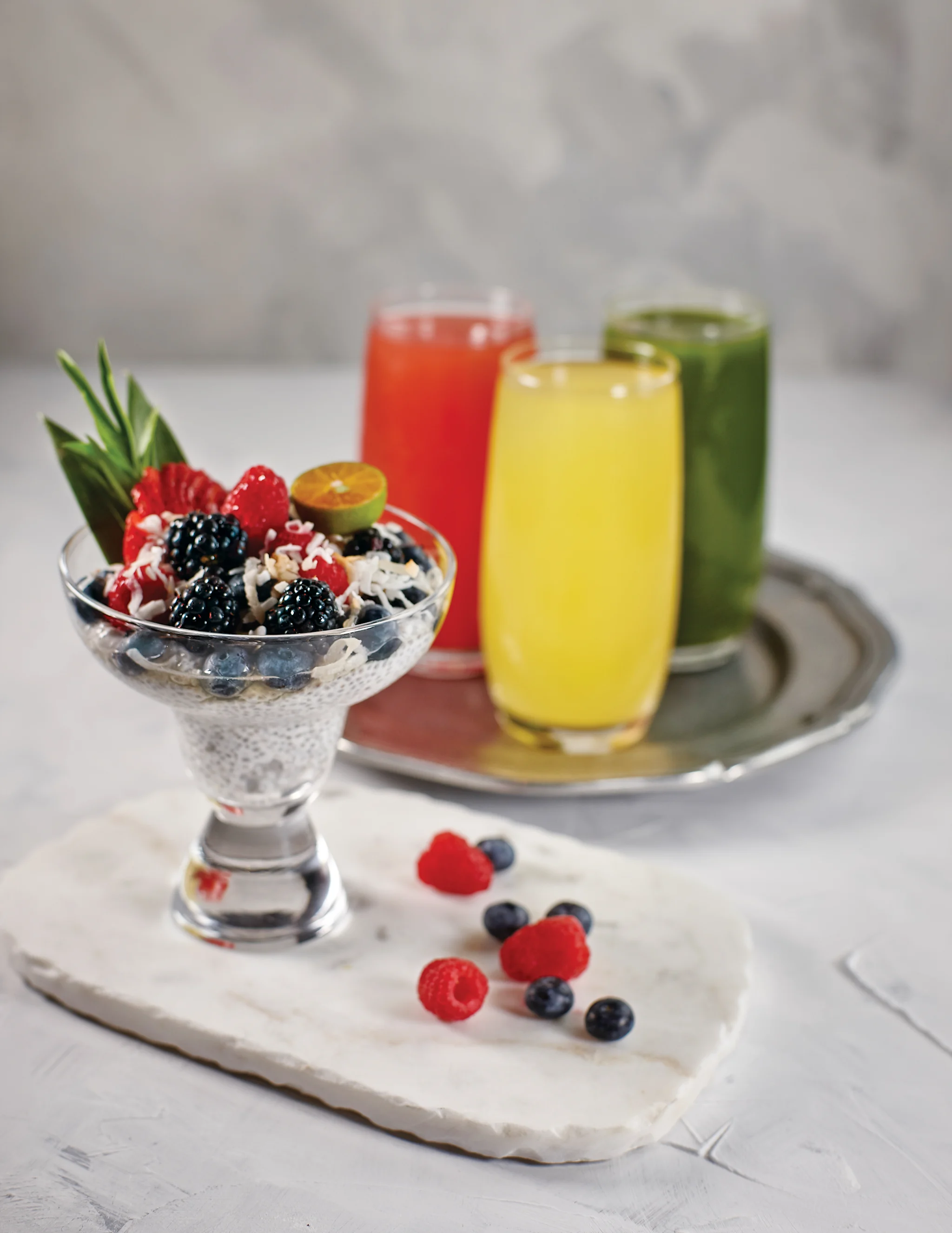 Chia pudding with berries and three colorful smoothies in red, yellow, and green on a tray.