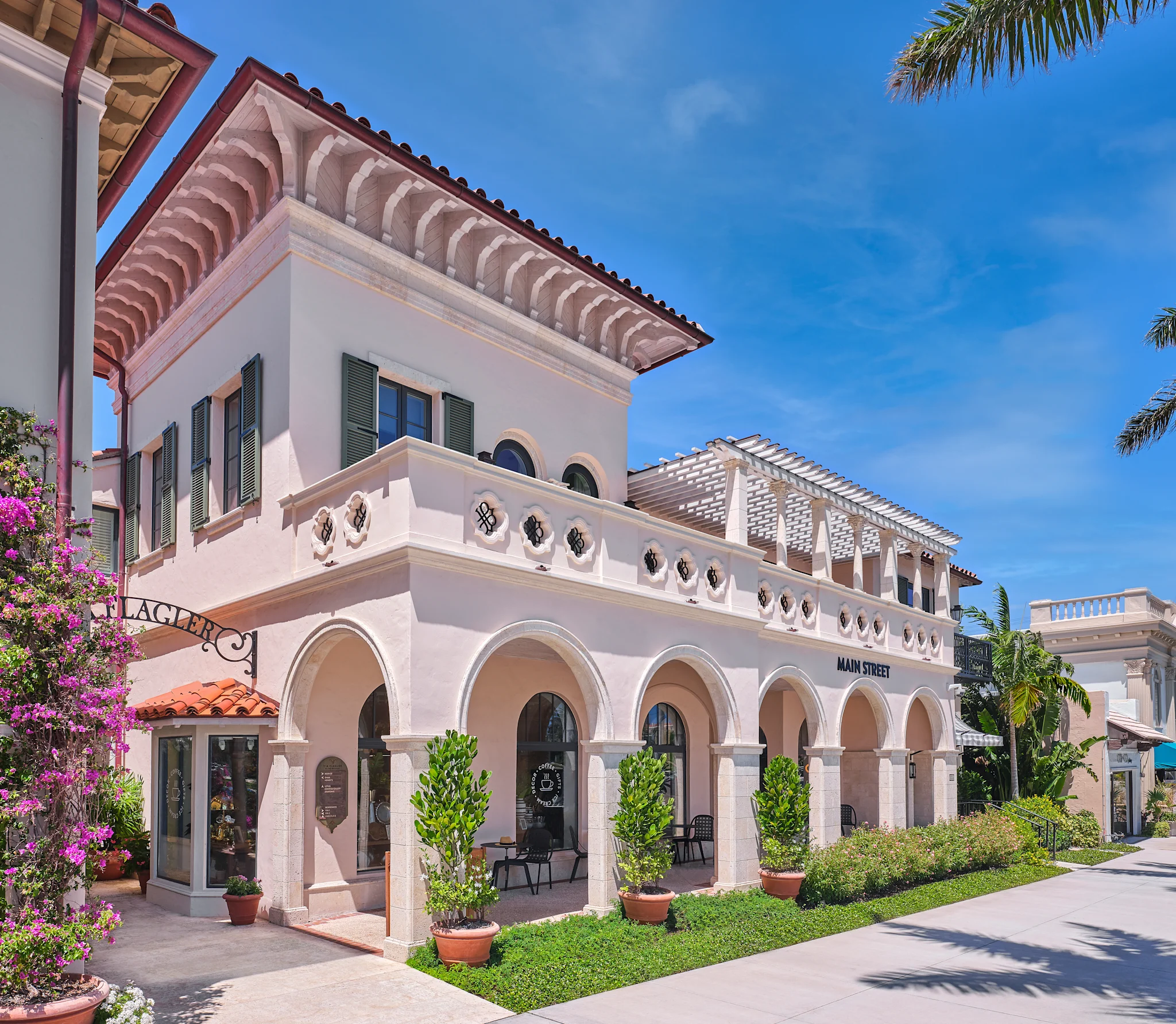 White Mediterranean-style building with arched colonnade, red tile roof, and palm trees under blue sky.