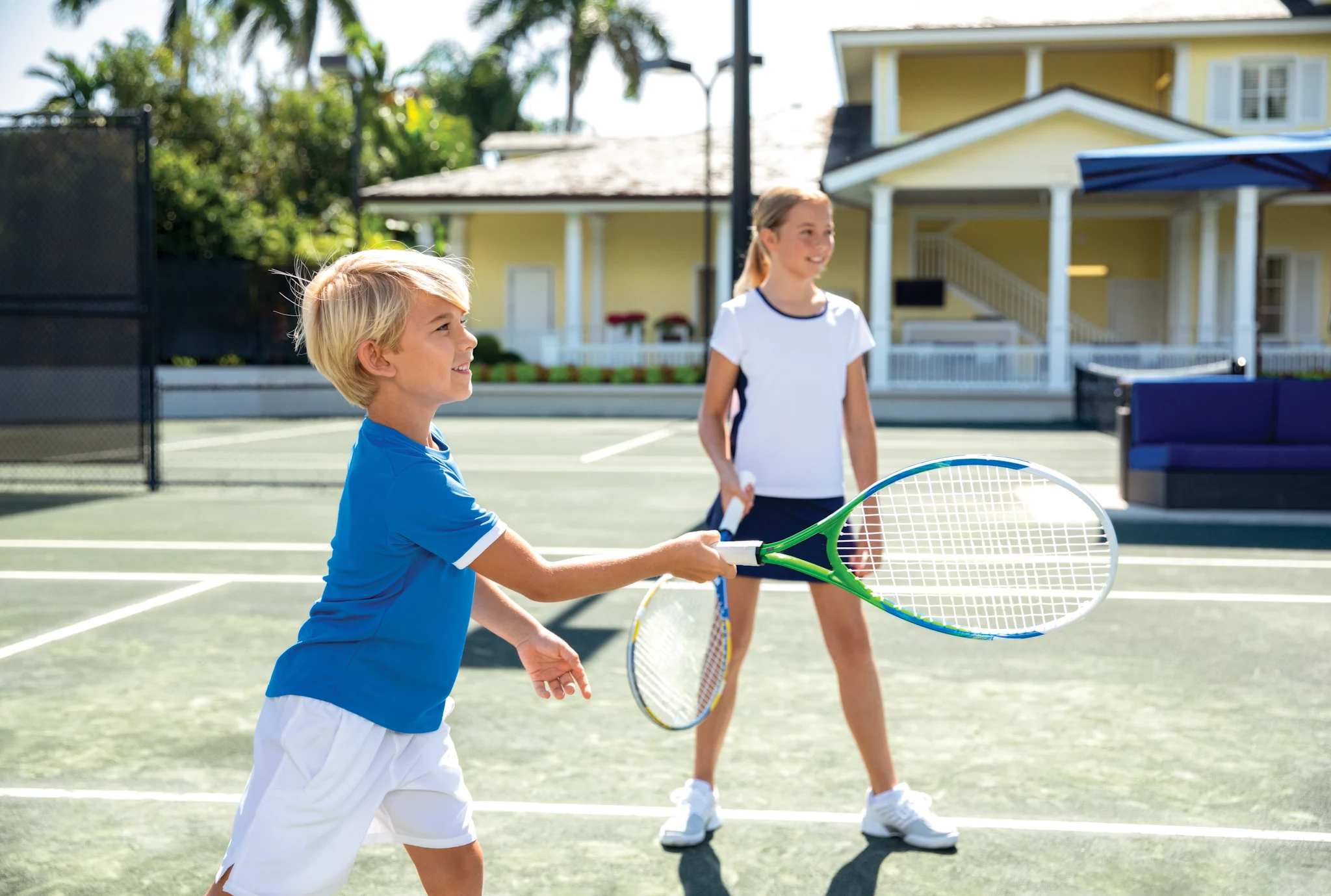Two children playing tennis on a sunny outdoor court with a yellow house in the background.